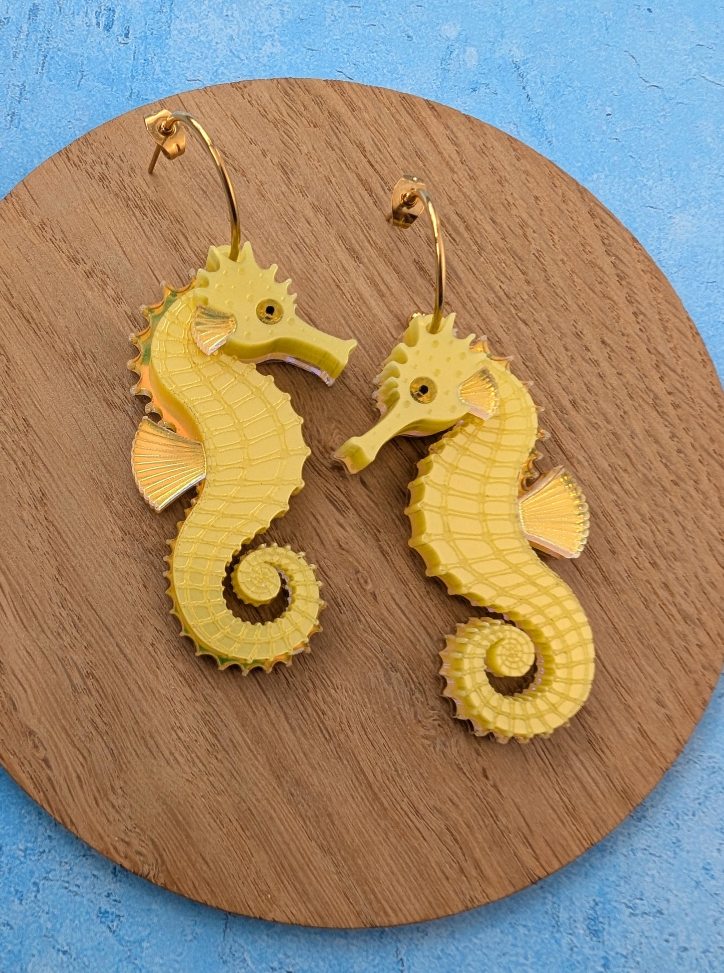 A pair of yellow seahorse-shaped dangle earrings with gold-tone hoops, displayed on a round wooden board against a textured blue background.