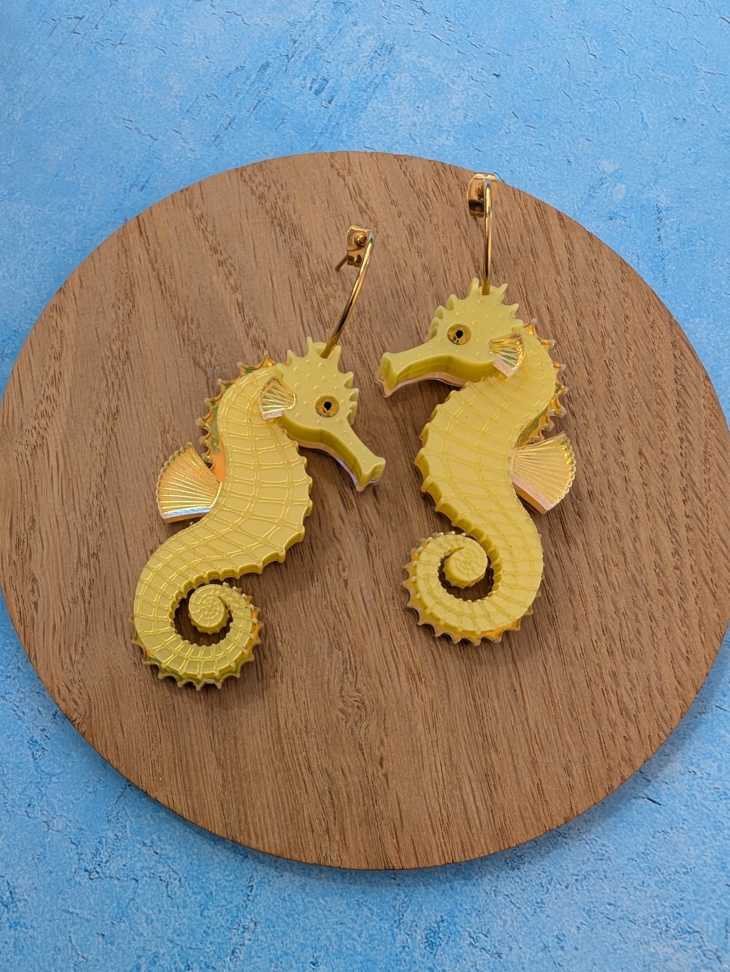 A pair of yellow seahorse-shaped earrings with gold hoops, displayed on a round wooden coaster against a textured blue background.