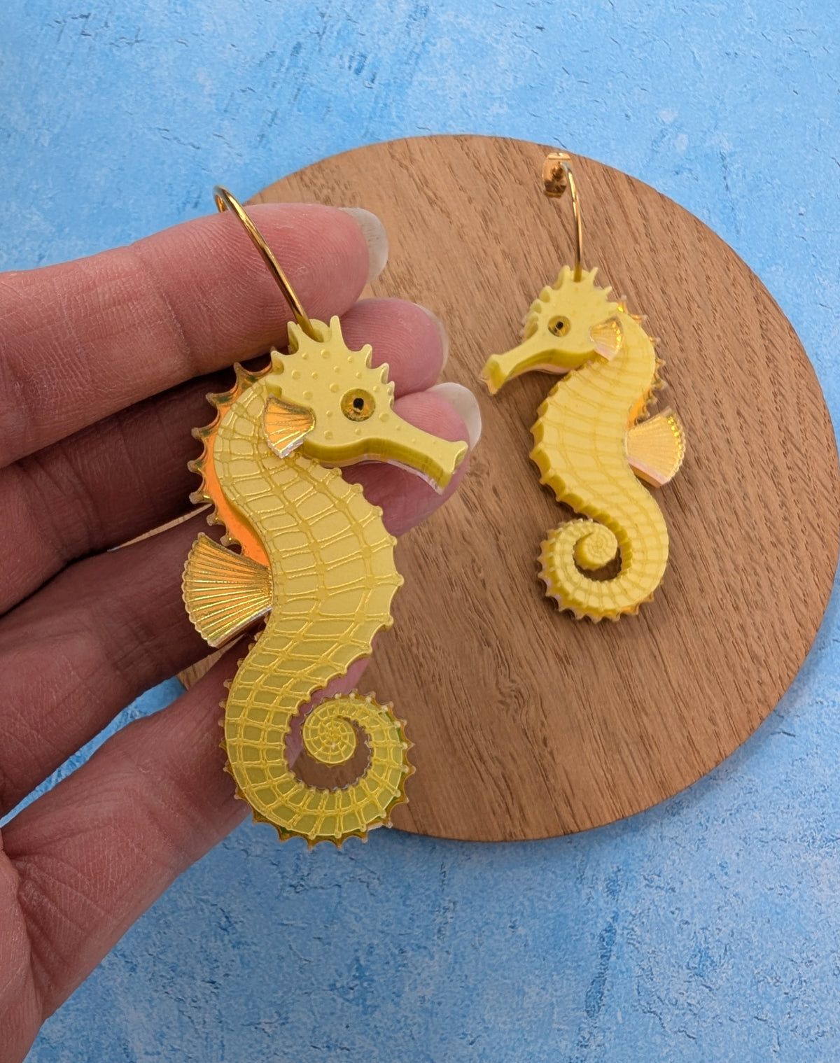 Yellow seahorse-shaped earrings held in a hand, with a matching pair resting on a round wooden coaster against a textured blue background.
