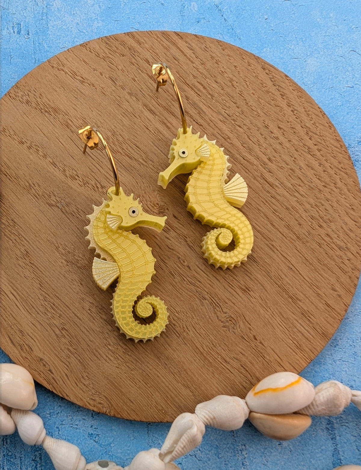 Yellow seahorse-shaped drop earrings with gold-tone hoops displayed on a round wooden board with seashells against a blue background.