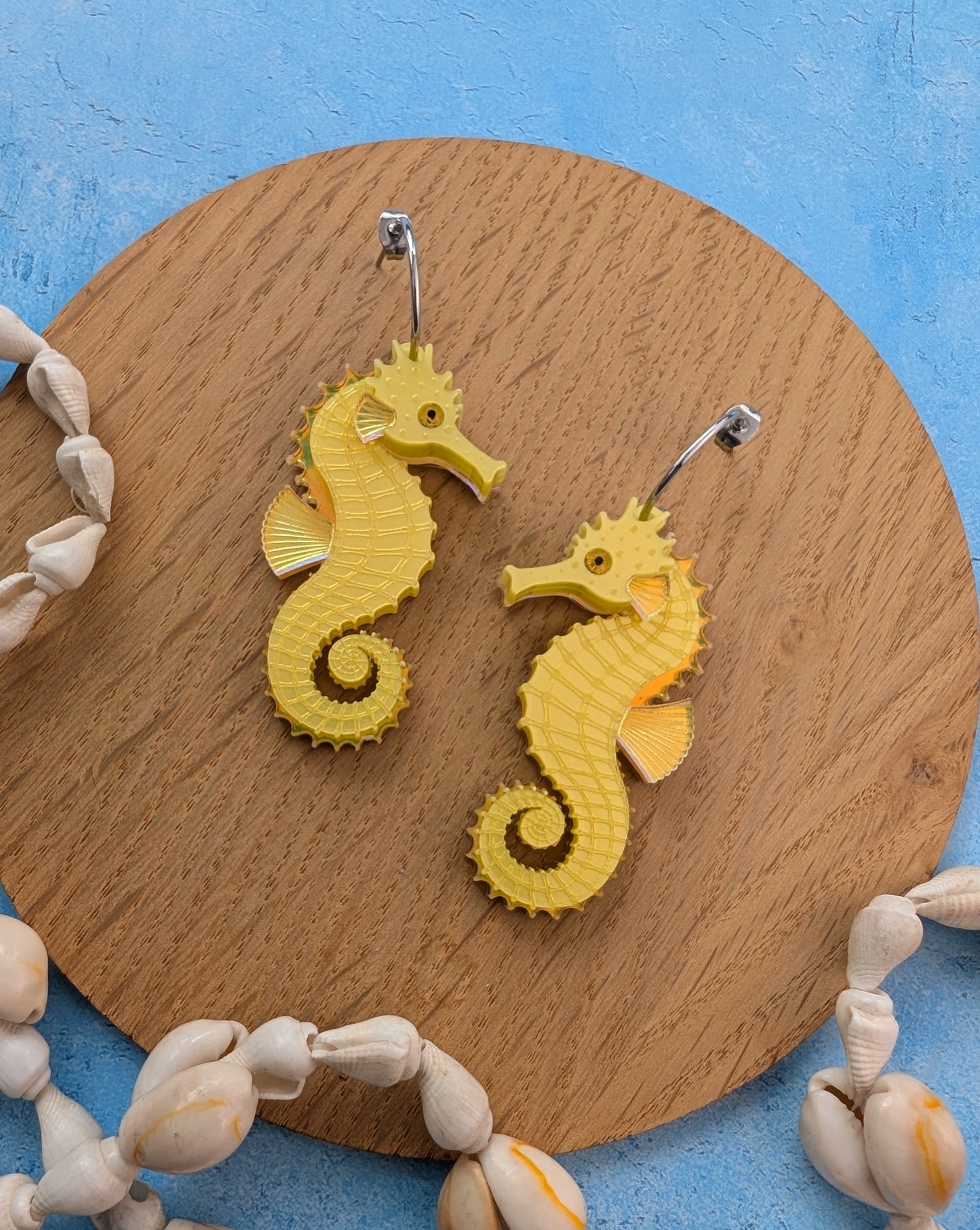 A pair of yellow seahorse-shaped earrings displayed on a round wooden board, surrounded by seashells on a textured blue background.