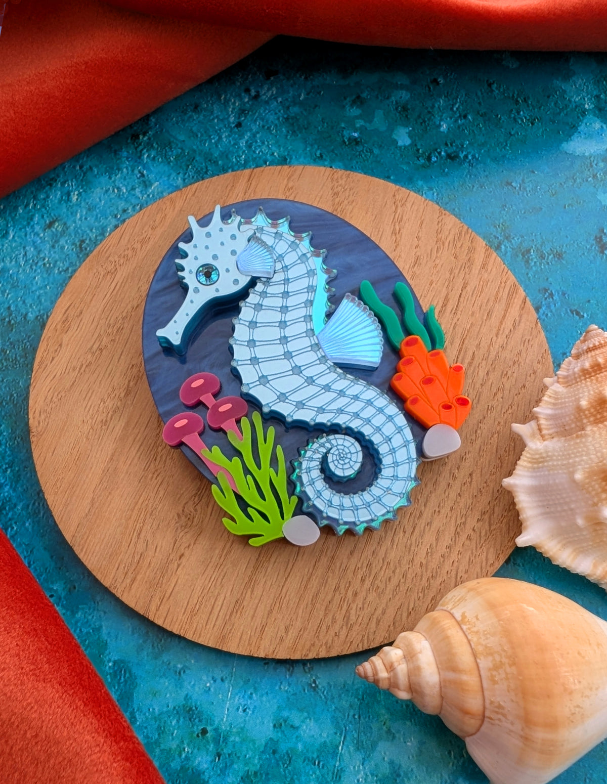 A handmade laser cut acrylic seahorse brooch displayed on a round wooden coaster, surrounded by seashells on a textured blue surface with a red fabric background.