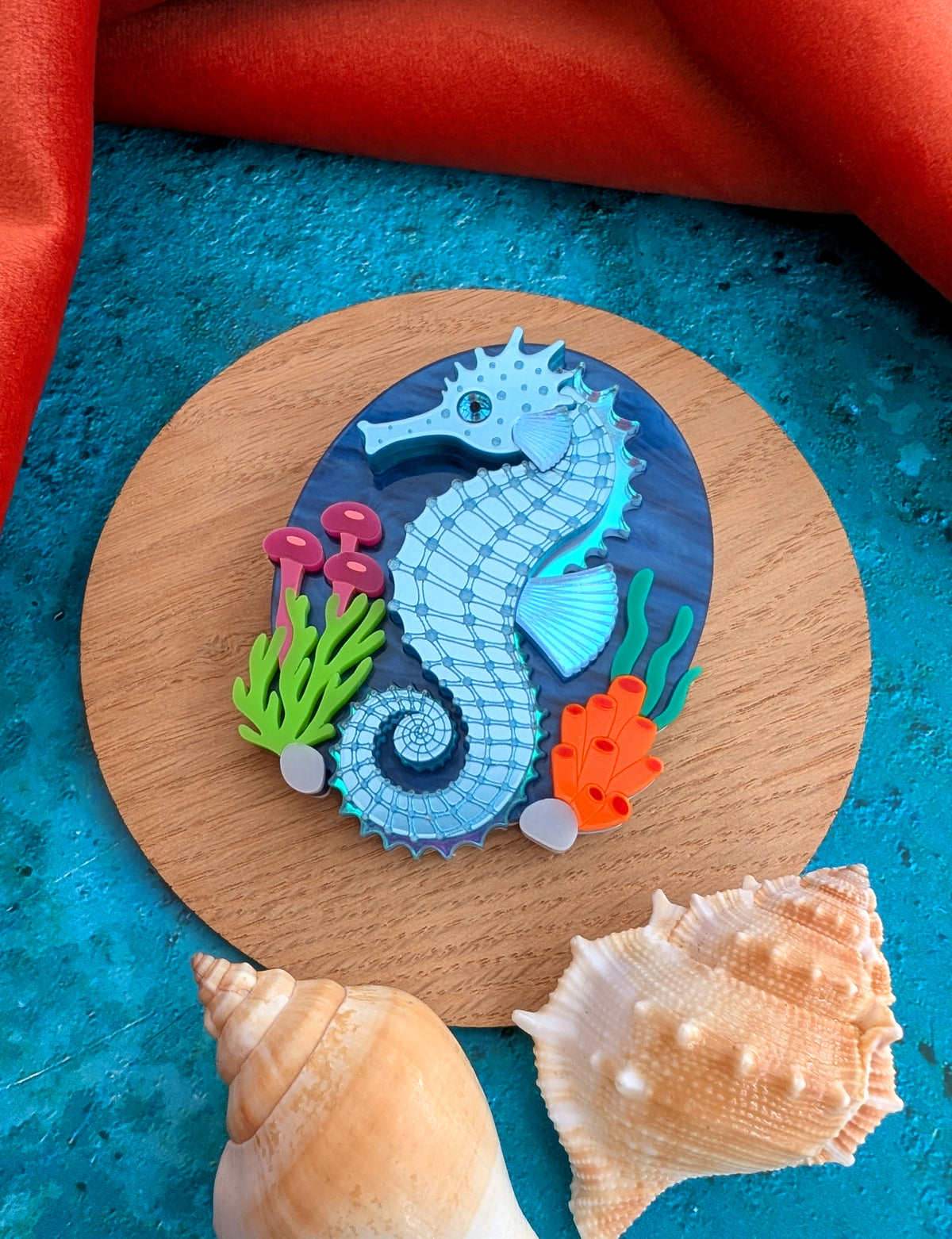 A handmade laser cut acrylic seahorse brooch displayed on a round wooden coaster, surrounded by seashells on a textured blue surface with a red fabric background.