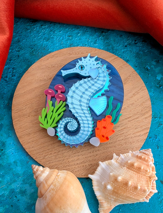 A handmade laser cut acrylic seahorse brooch displayed on a round wooden coaster, surrounded by seashells on a textured blue surface with a red fabric background.