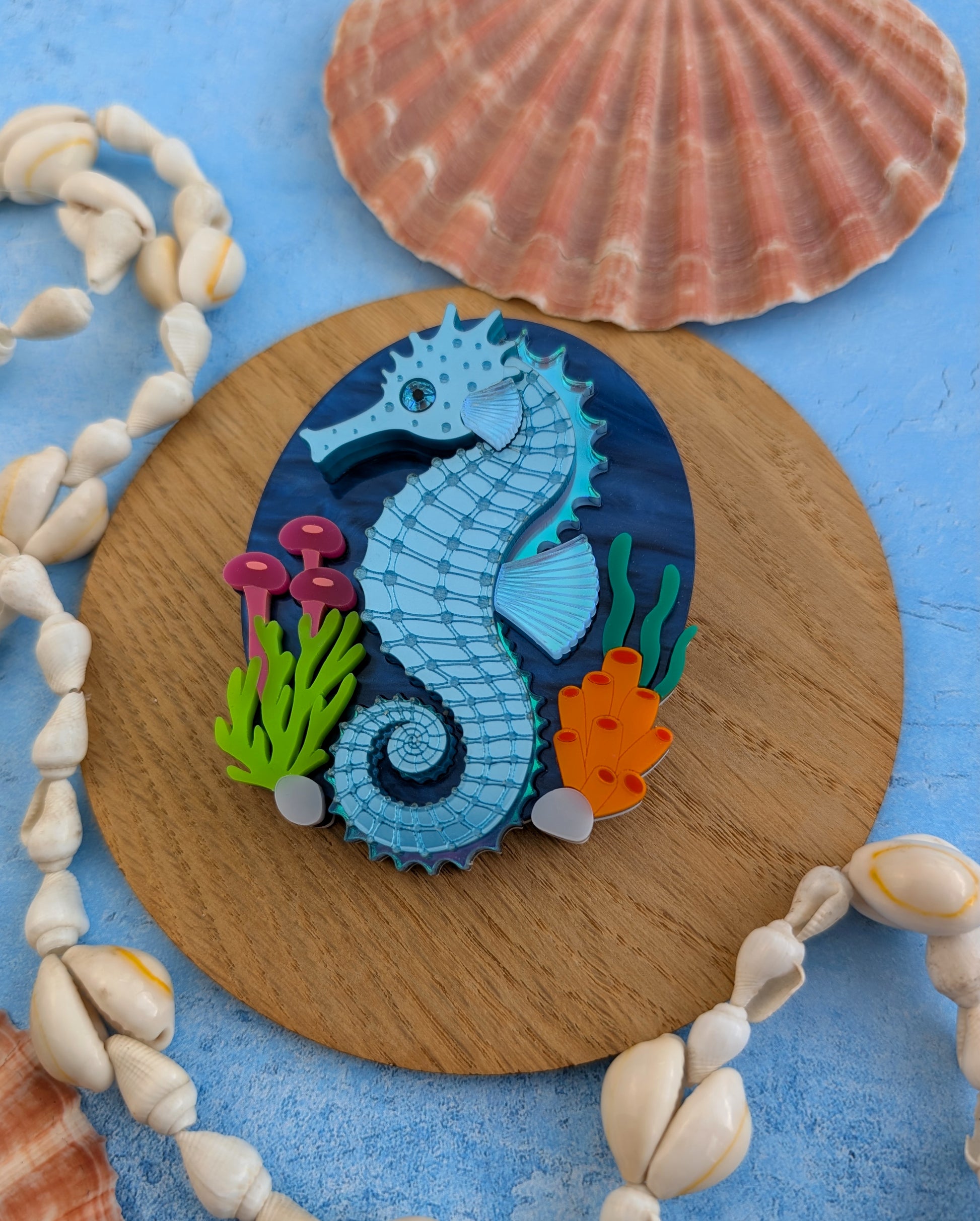 A handmade laser cut acrylic seahorse brooch displayed on a round wooden coaster, surrounded by seashells on a textured blue surface with a red fabric background.