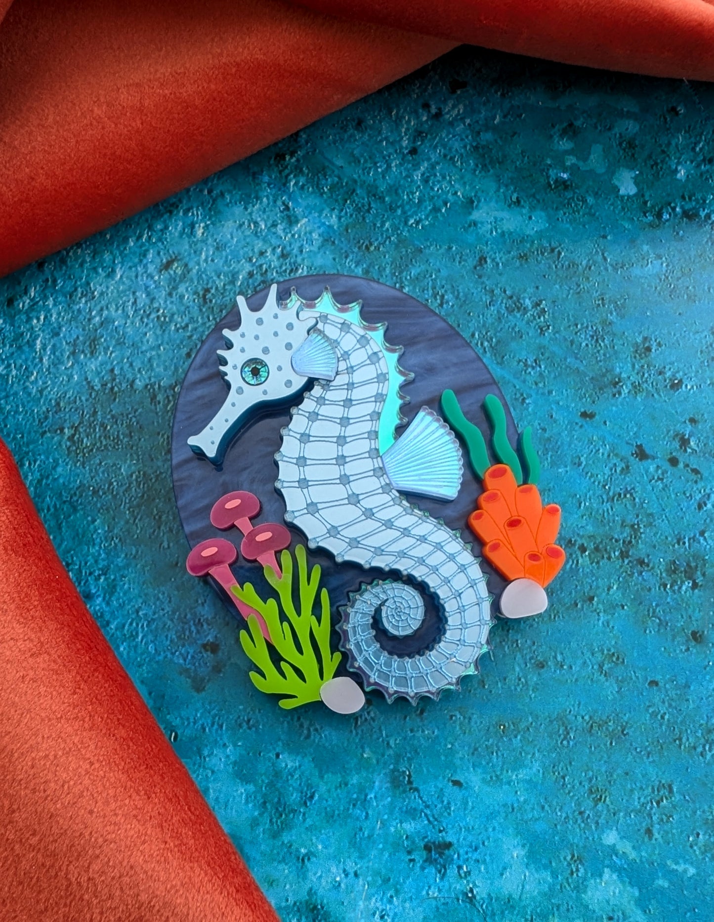 A handmade laser cut acrylic seahorse brooch displayed on a round wooden coaster, surrounded by seashells on a textured blue surface with a red fabric background.