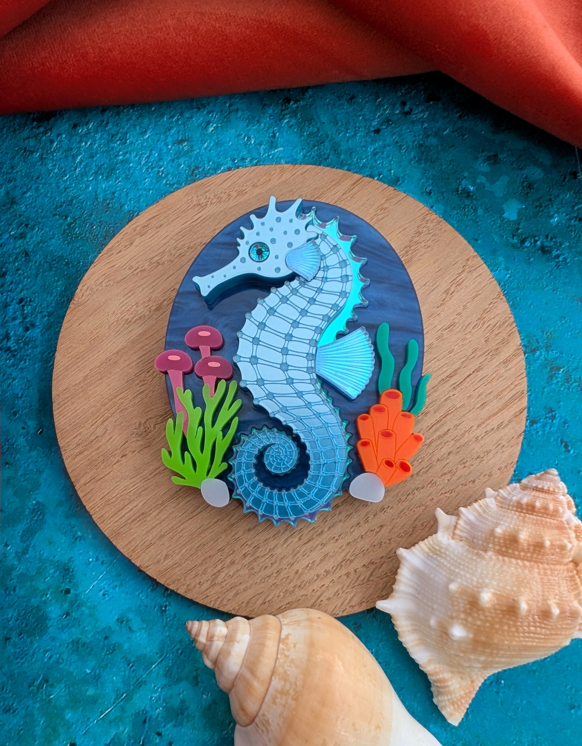 A handmade laser cut acrylic seahorse brooch displayed on a round wooden coaster, surrounded by seashells on a textured blue surface with a red fabric background.