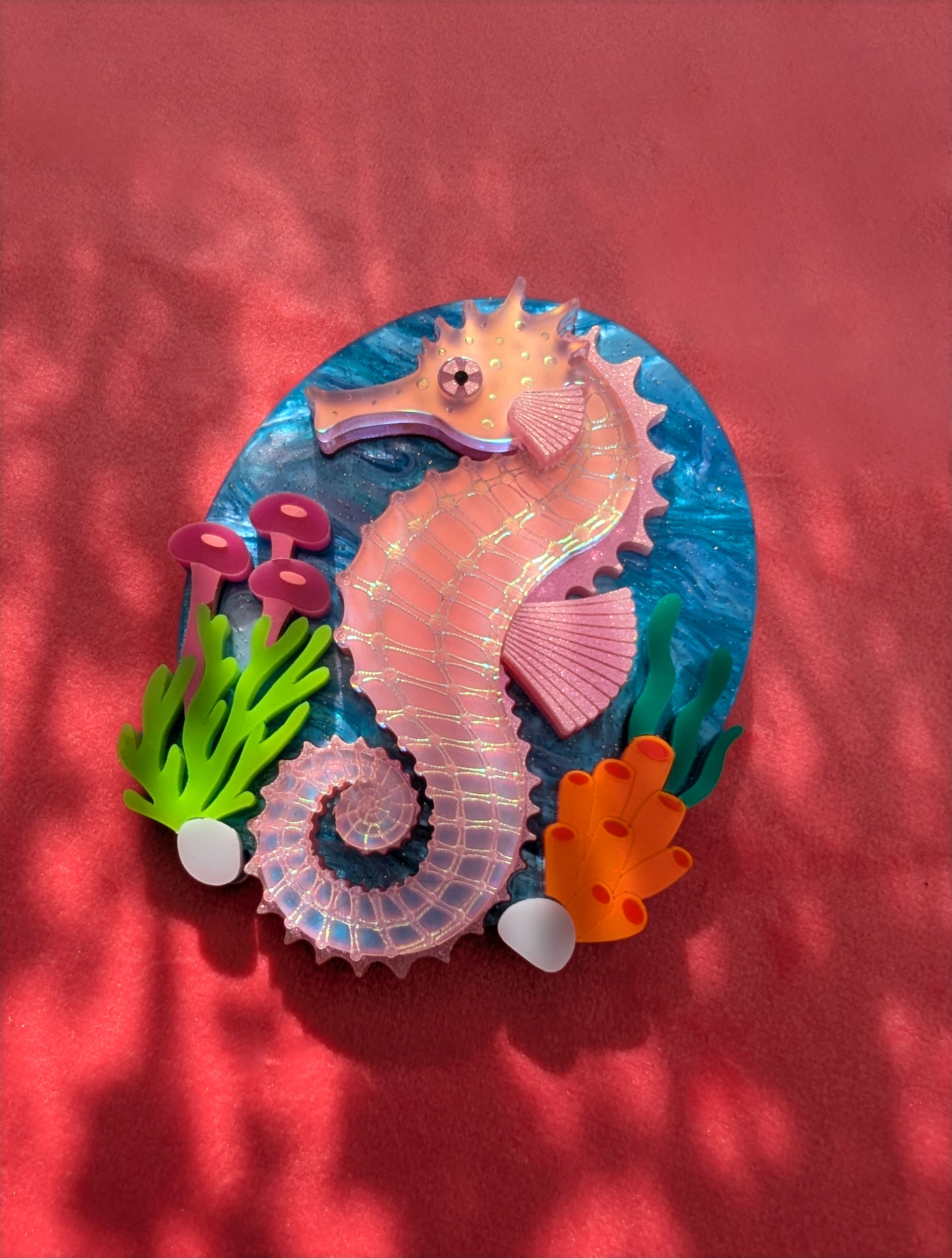 A handmade laser cut acrylic iridescent pink seahorse brooch with a blue sea background and sea plants/coral