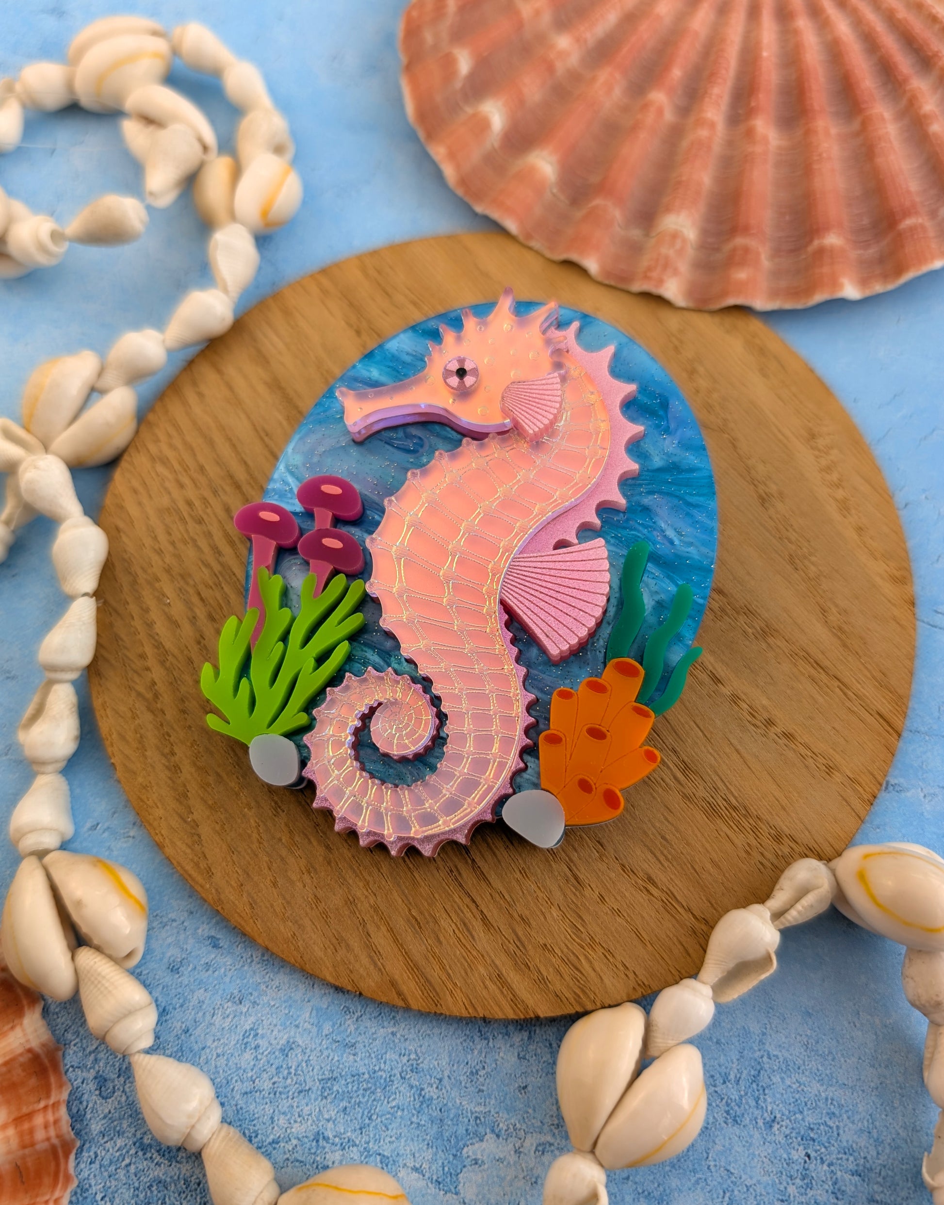 A handmade laser cut acrylic iridescent pink seahorse brooch with a blue sea background and sea plants/coral