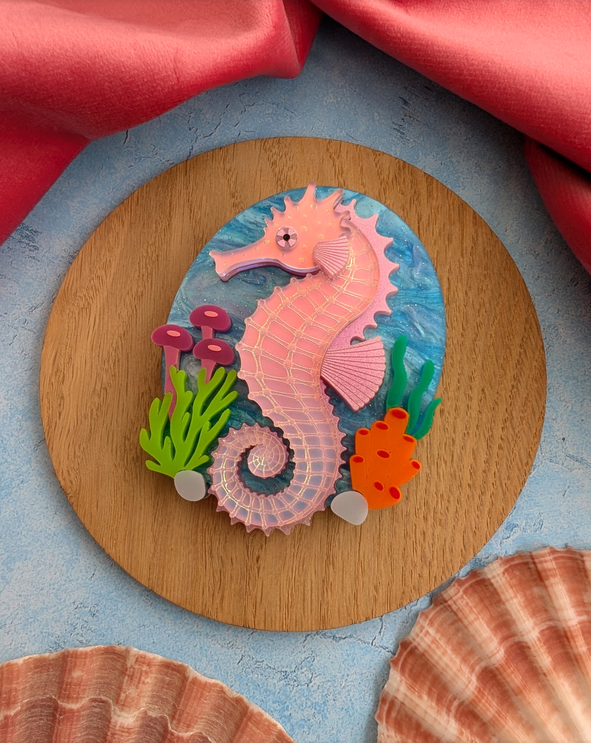 A handmade laser cut acrylic iridescent pink seahorse brooch with a blue sea background and sea plants/coral