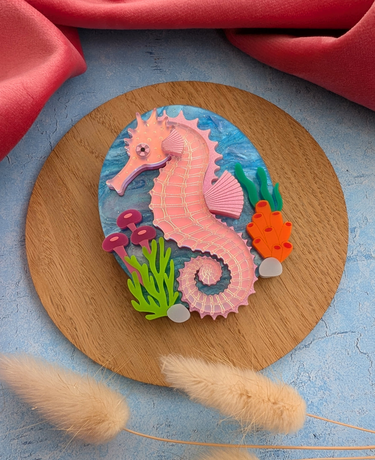 A handmade laser cut acrylic iridescent pink seahorse brooch with a blue sea background and sea plants/coral