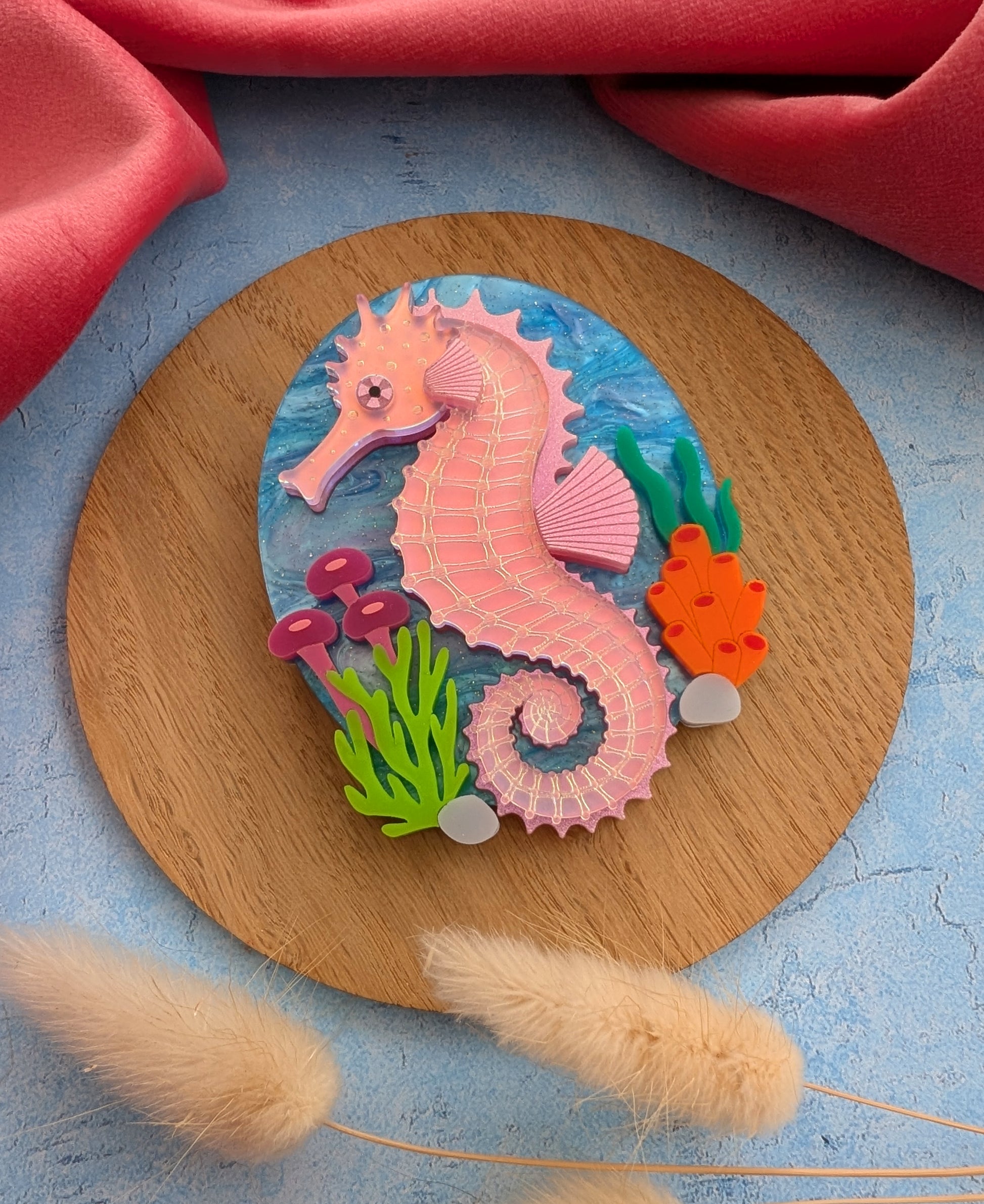A handmade laser cut acrylic iridescent pink seahorse brooch with a blue sea background and sea plants/coral