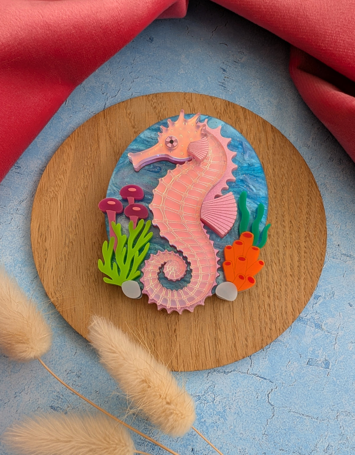 A handmade laser cut acrylic iridescent pink seahorse brooch with a blue sea background and sea plants/coral