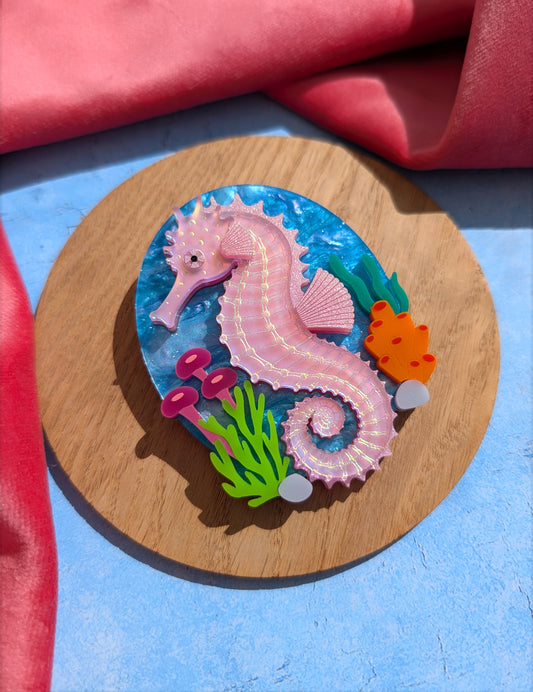 A handmade laser cut acrylic iridescent pink seahorse brooch with a blue sea background and sea plants/coral