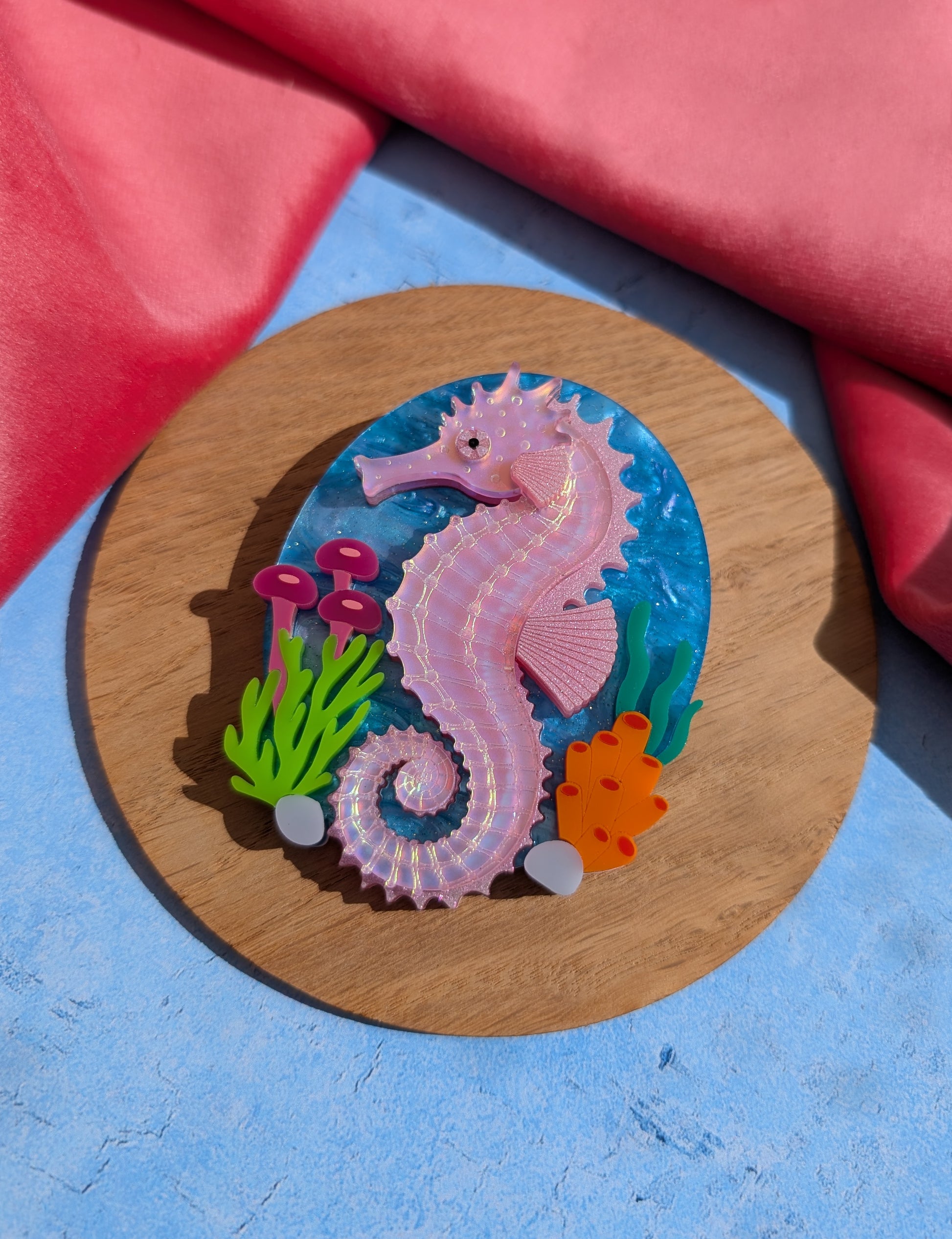 A handmade laser cut acrylic iridescent pink seahorse brooch with a blue sea background and sea plants/coral