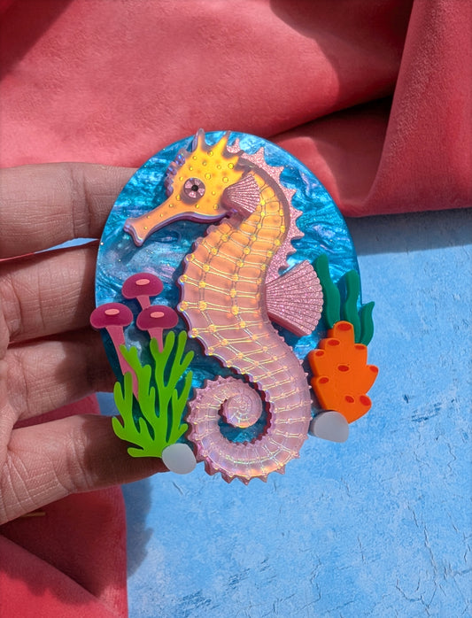 A handmade laser cut acrylic iridescent pink seahorse brooch with a blue sea background and sea plants/coral