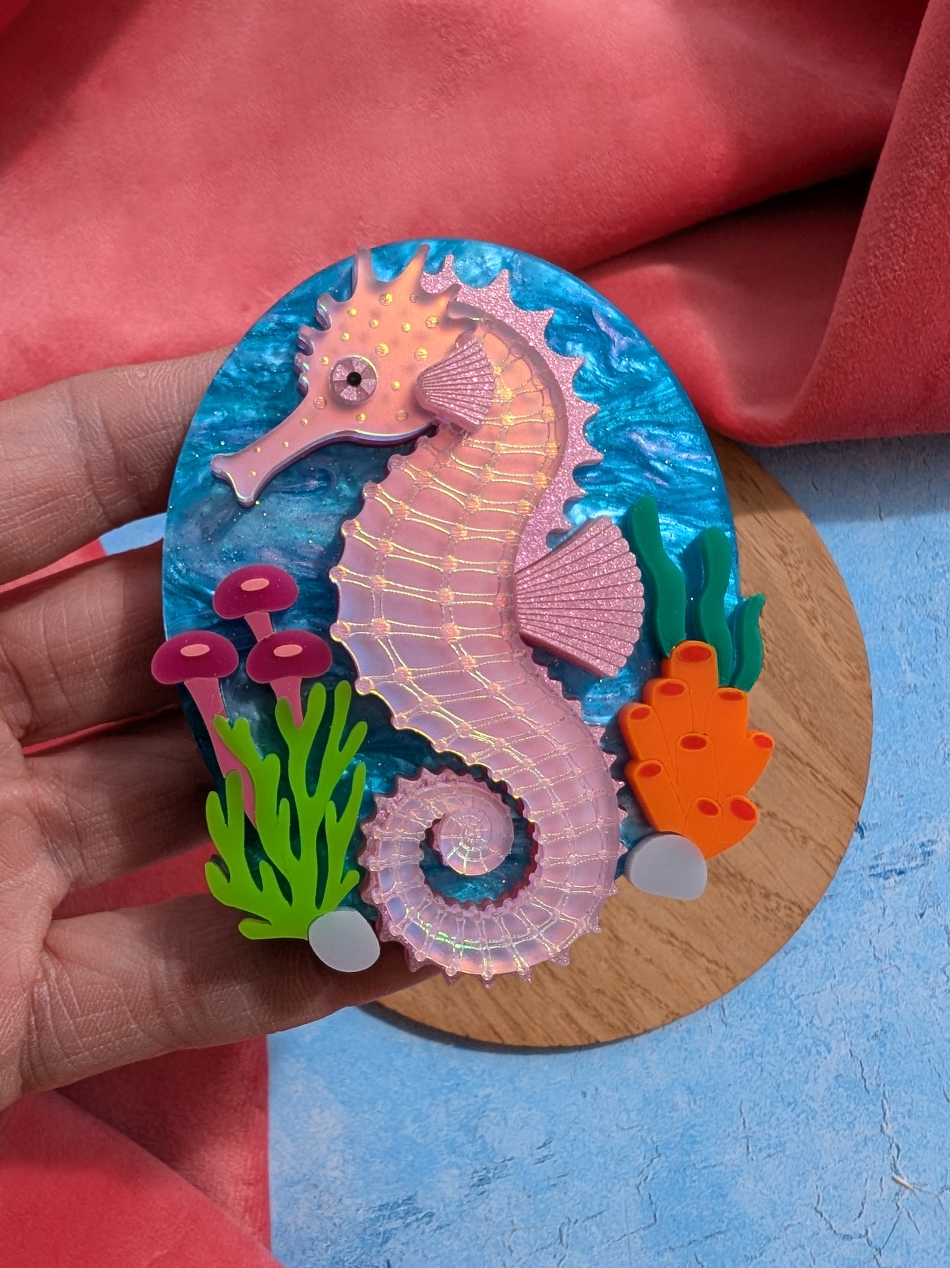 A handmade laser cut acrylic iridescent pink seahorse brooch with a blue sea background and sea plants/coral