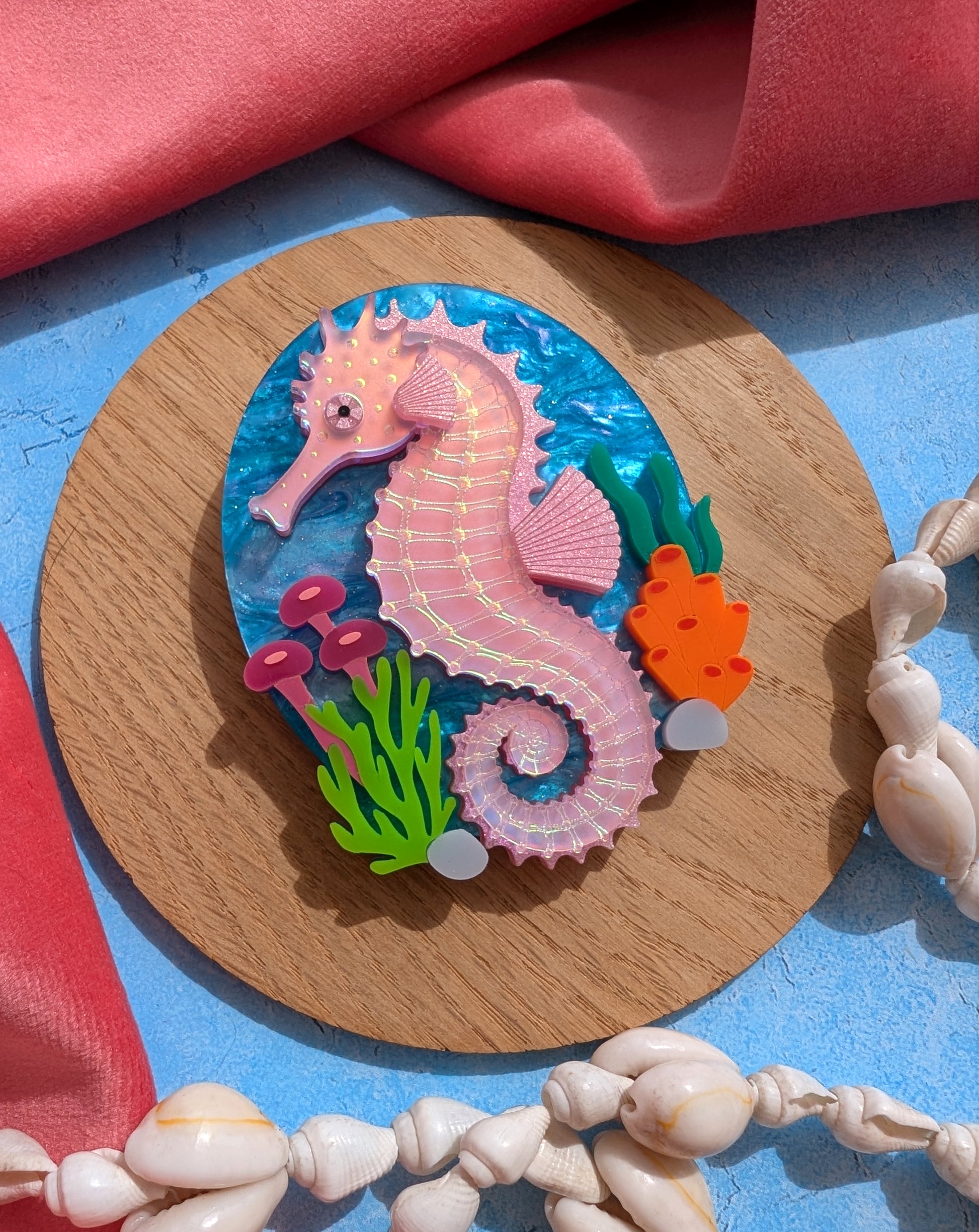 A handmade laser cut acrylic iridescent pink seahorse brooch with a blue sea background and sea plants/coral