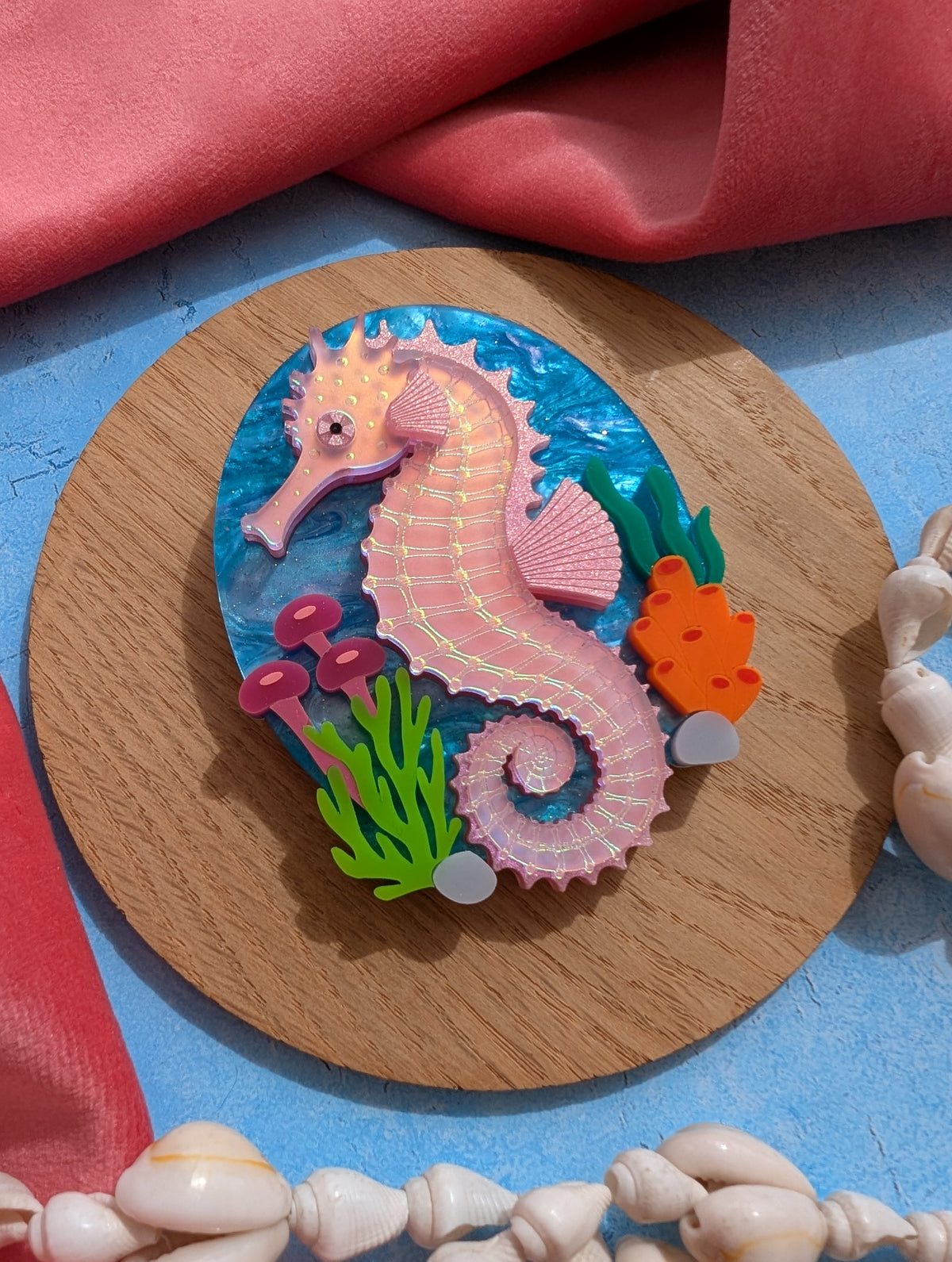 A handmade laser cut acrylic iridescent pink seahorse brooch with a blue sea background and sea plants/coral
