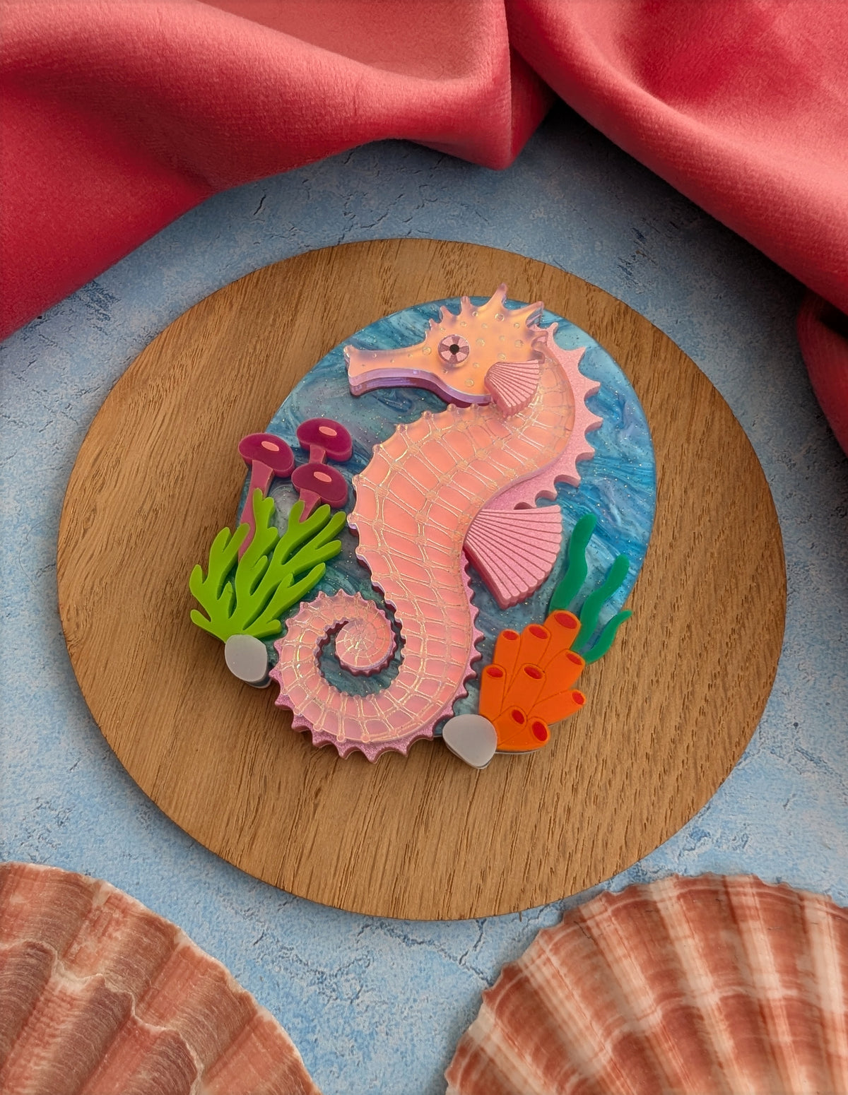 A handmade laser cut acrylic iridescent pink seahorse brooch with a blue sea background and sea plants/coral