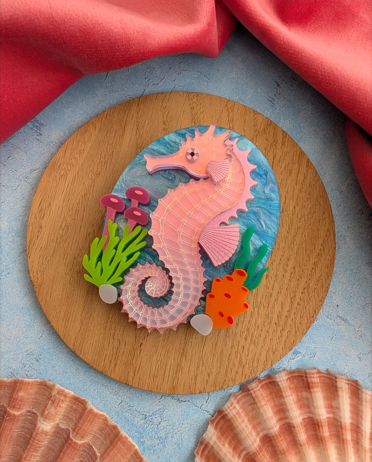 A handmade laser cut acrylic iridescent pink seahorse brooch with a blue sea background and sea plants/coral