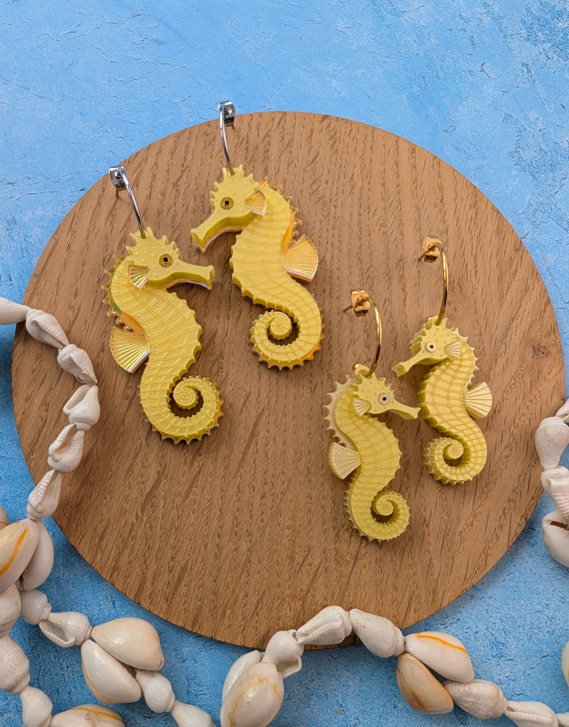Two pairs of seahorse-shaped earrings displayed on a round wooden board, surrounded by seashells on a textured blue background.