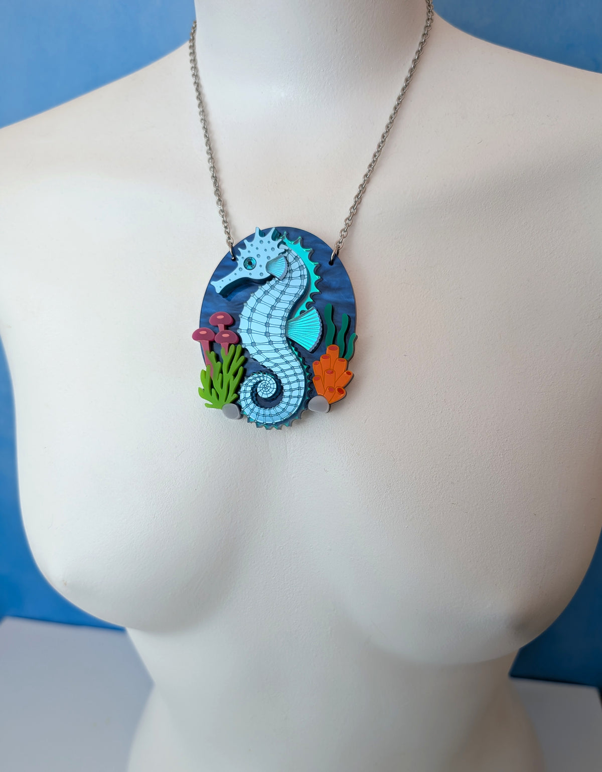 A laser cut handmade acrylic necklace with an oval pendant featuring a blue and turquoise seahorse surrounded by colourful sea plants, displayed on a white mannequin torso against a blue background.