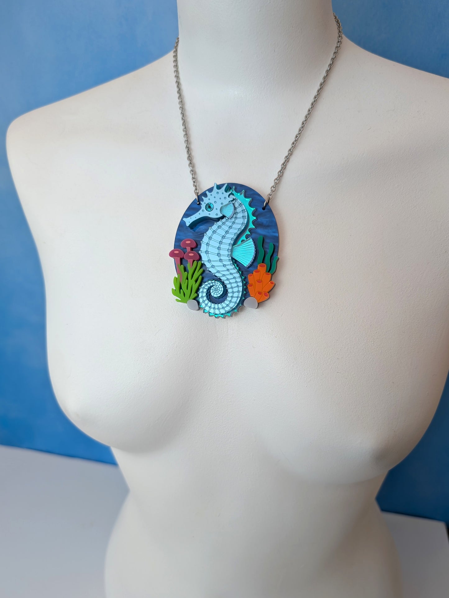 A laser cut handmade acrylic necklace with an oval pendant featuring a blue and turquoise seahorse surrounded by colourful sea plants, displayed on a white mannequin torso against a blue background.