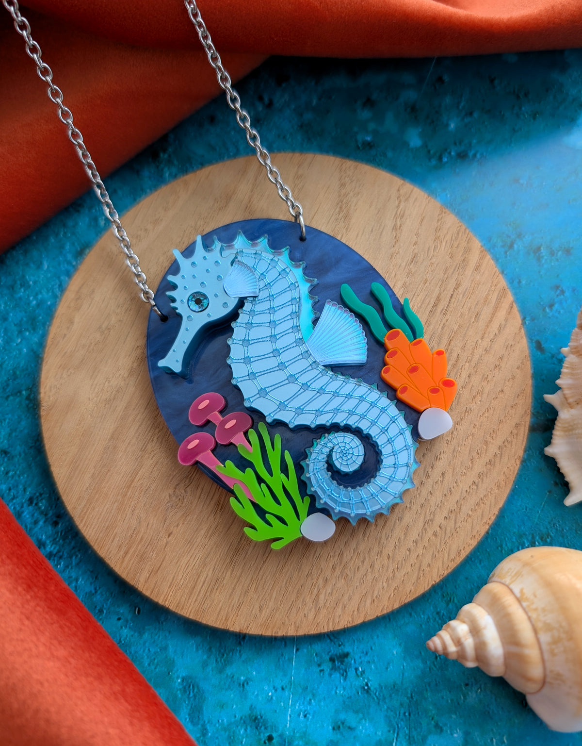 A laser cut handmade acrylic necklace with an oval pendant featuring a blue and turquoise seahorse surrounded by colourful sea plants, displayed on a white mannequin torso against a blue background.