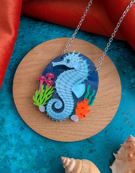 A laser cut handmade acrylic necklace with an oval pendant featuring a blue and turquoise seahorse surrounded by colourful sea plants, displayed on a white mannequin torso against a blue background.