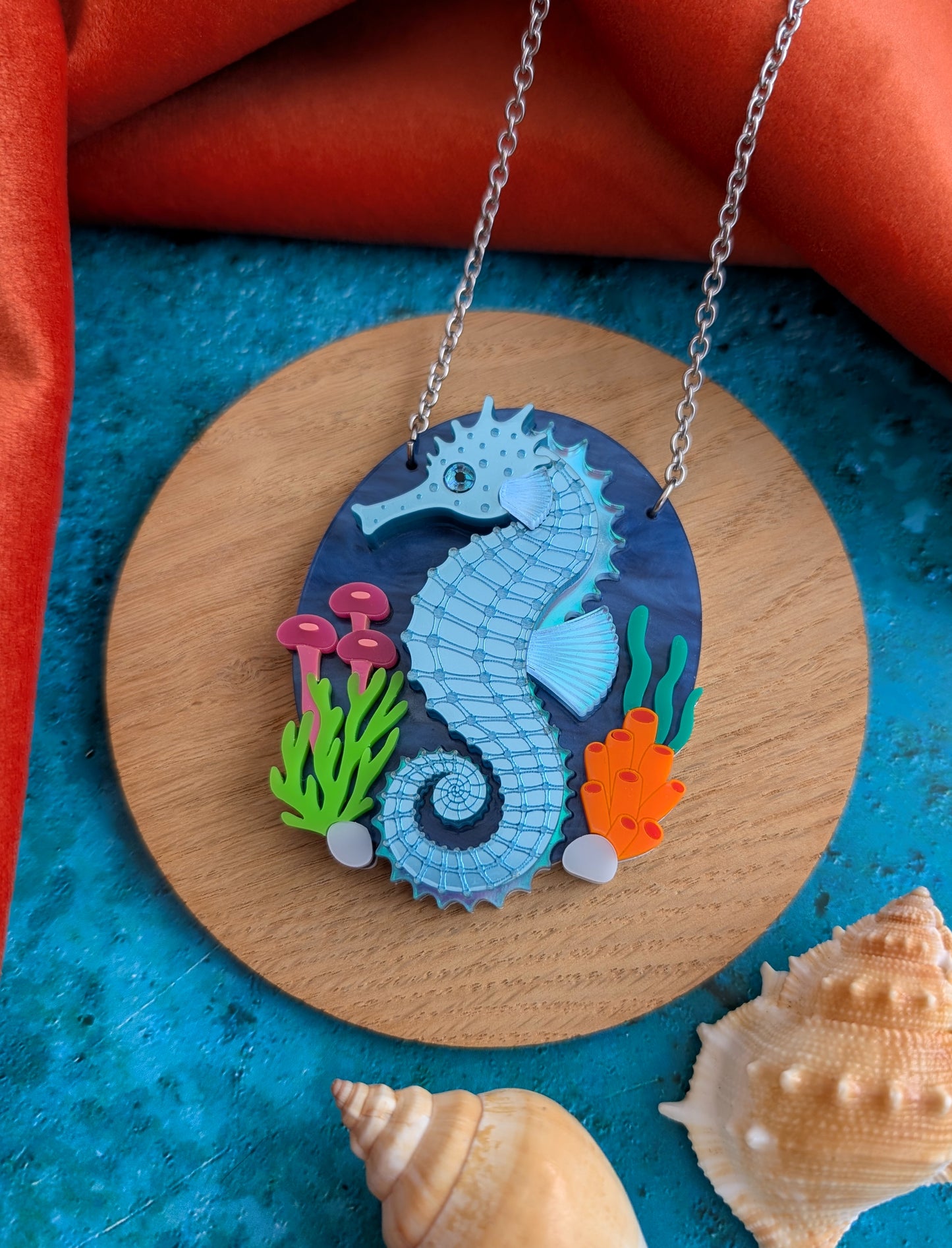 A laser cut handmade acrylic necklace with an oval pendant featuring a blue and turquoise seahorse surrounded by colourful sea plants, displayed on a white mannequin torso against a blue background.