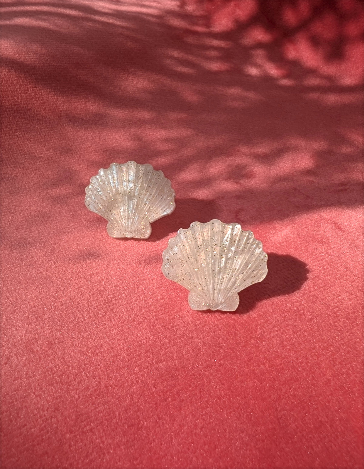 Two small laser cut acrylic shell stud earrings in a ivory pearl marble acrylic displayed on a textured pink surface with dappled shadows.