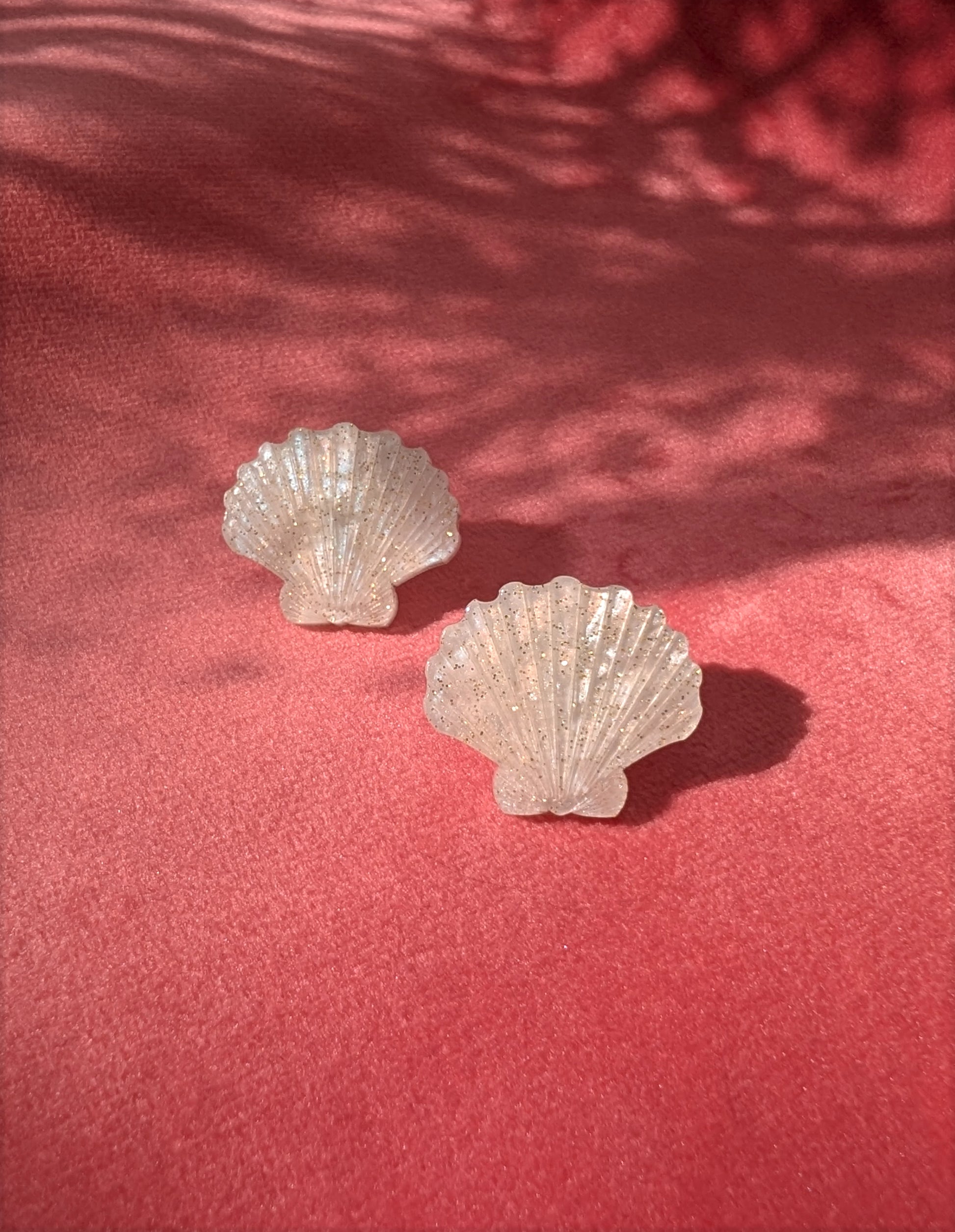 Two small laser cut acrylic shell stud earrings in a ivory pearl marble acrylic displayed on a textured pink surface with dappled shadows.