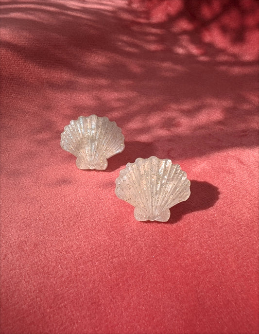 Two small laser cut acrylic shell stud earrings in a ivory pearl marble acrylic displayed on a textured pink surface with dappled shadows.