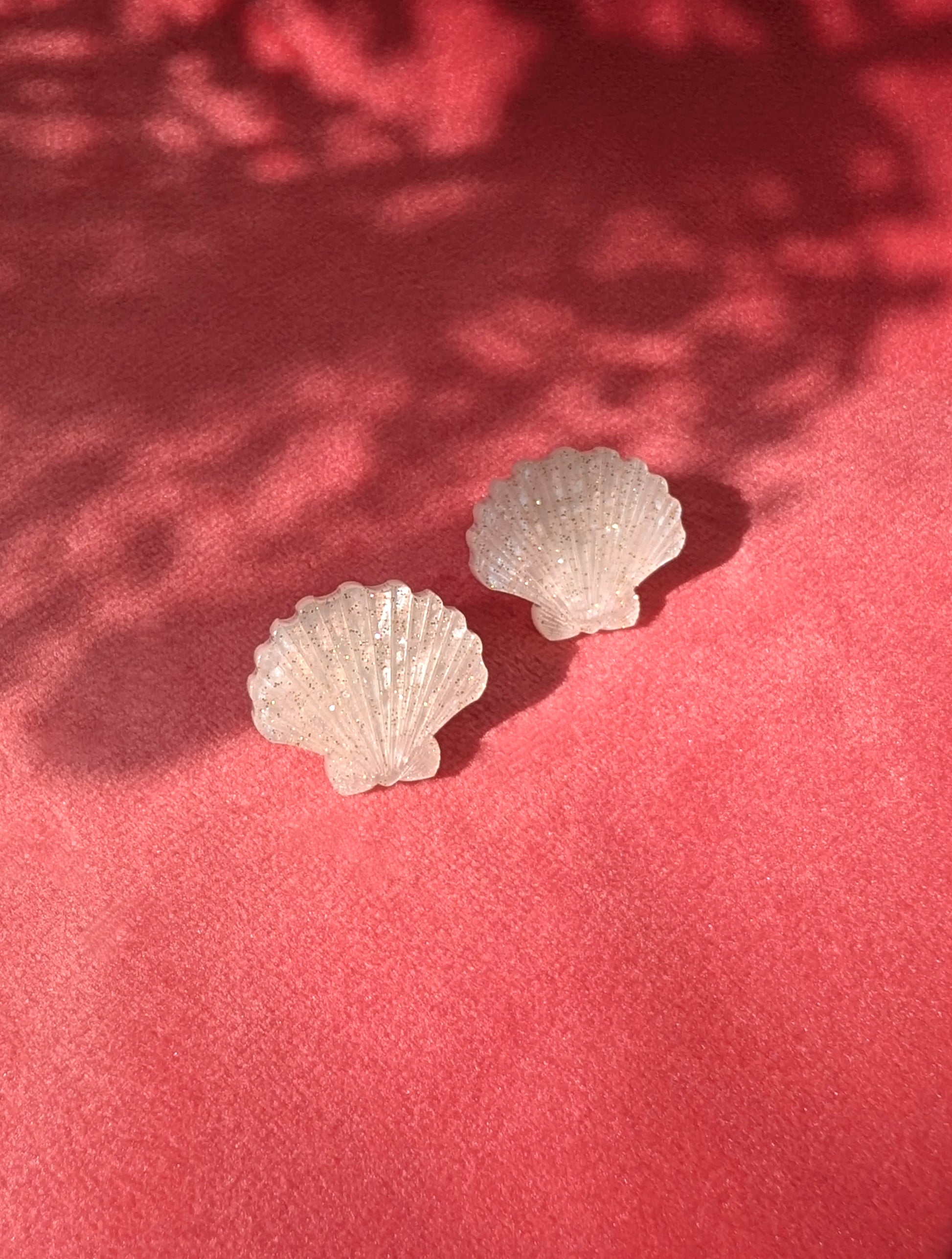 Two small laser cut acrylic shell stud earrings in a ivory pearl marble acrylic displayed on a textured pink surface with dappled shadows.