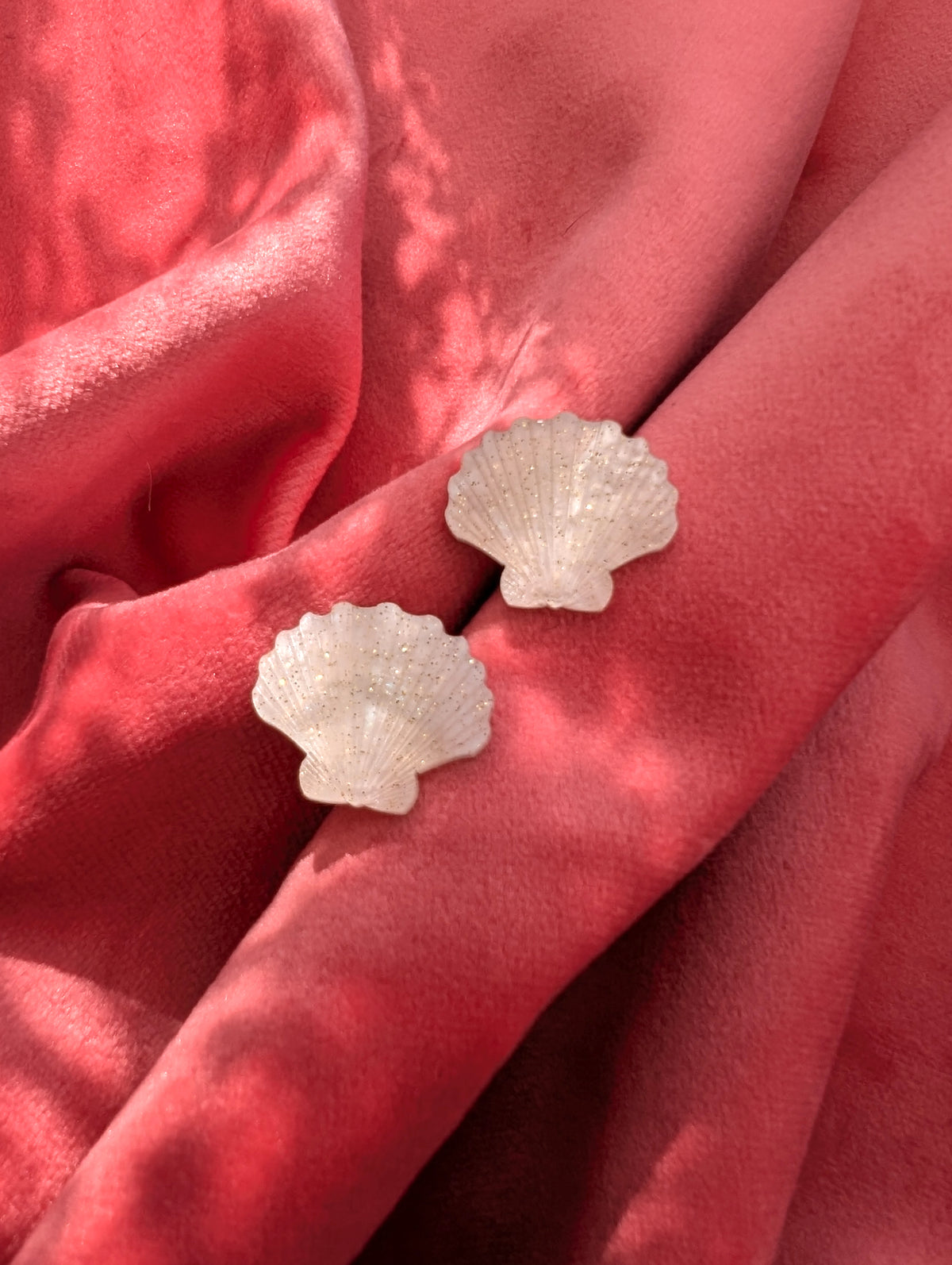 Two small laser cut acrylic shell stud earrings in a ivory pearl marble acrylic displayed on a textured pink surface with dappled shadows.
