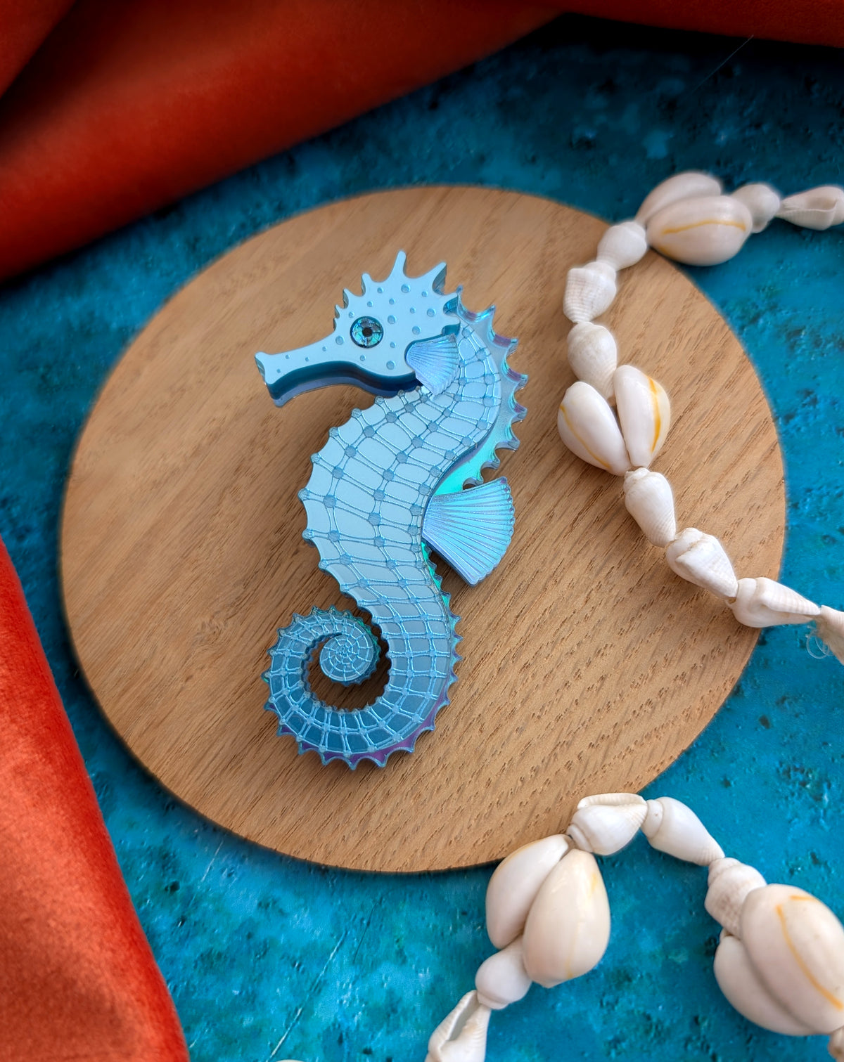 A light blue seahorse-shaped brooch with a textured, layered design, displayed on a round wooden coaster. The coaster is set against a textured blue background with decorative white seashells and draped orange fabric.