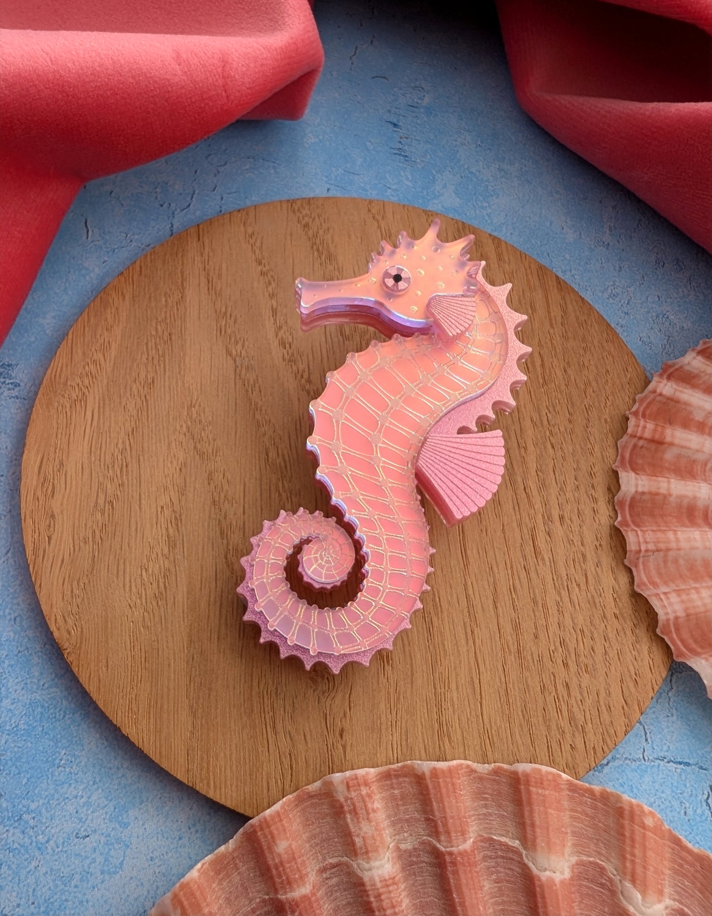 A pink acrylic seahorse brooch displayed on a round wooden board, surrounded by seashells and coral-coloured fabric on a blue textured surface.