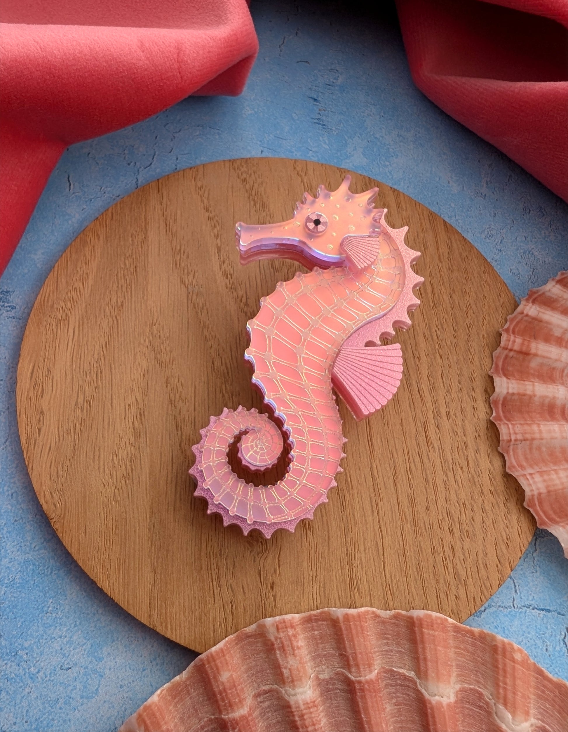 A pink acrylic seahorse brooch displayed on a round wooden board, surrounded by seashells and coral-coloured fabric on a blue textured surface.