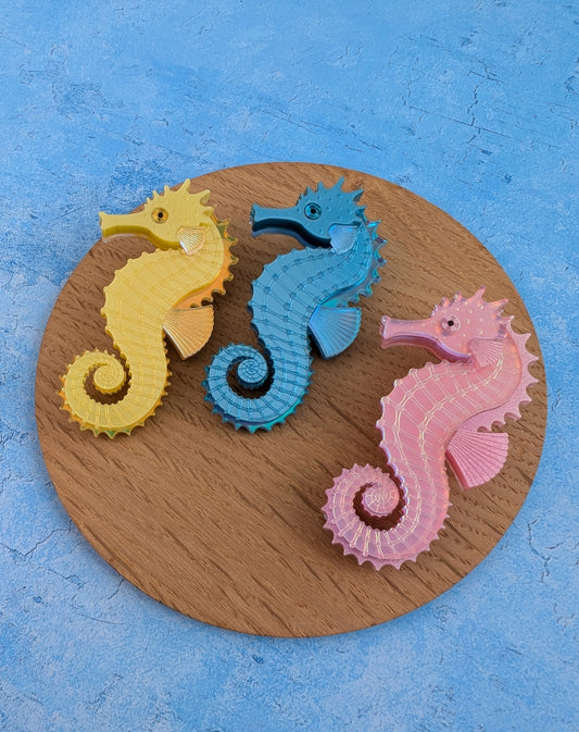 Three handmade acrylic seahorse brooches in yellow, blue, and pink, arranged on a round wooden board against a textured blue background.