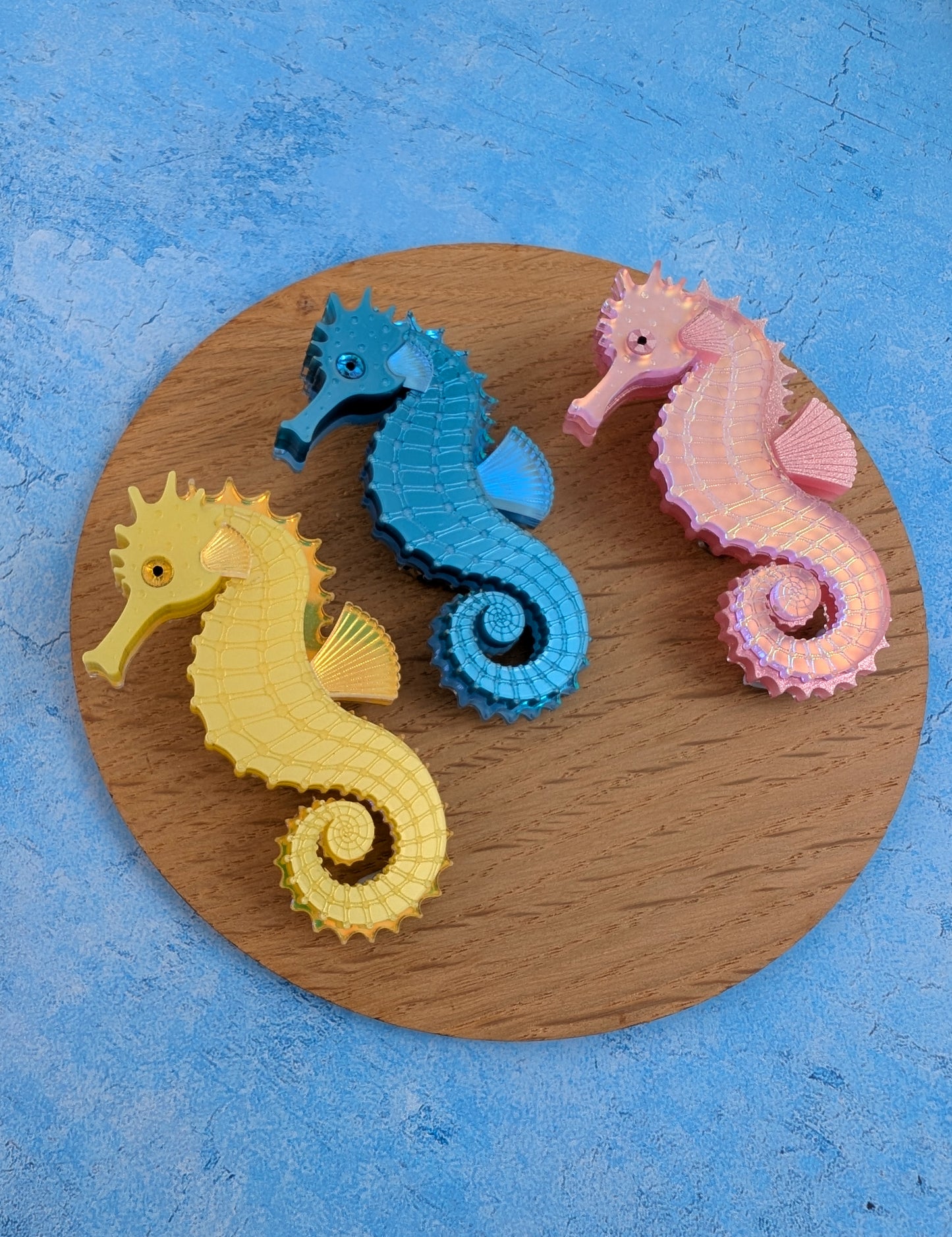 Three handmade acrylic seahorse brooches in yellow, blue, and pink, arranged on a round wooden board against a textured blue background.