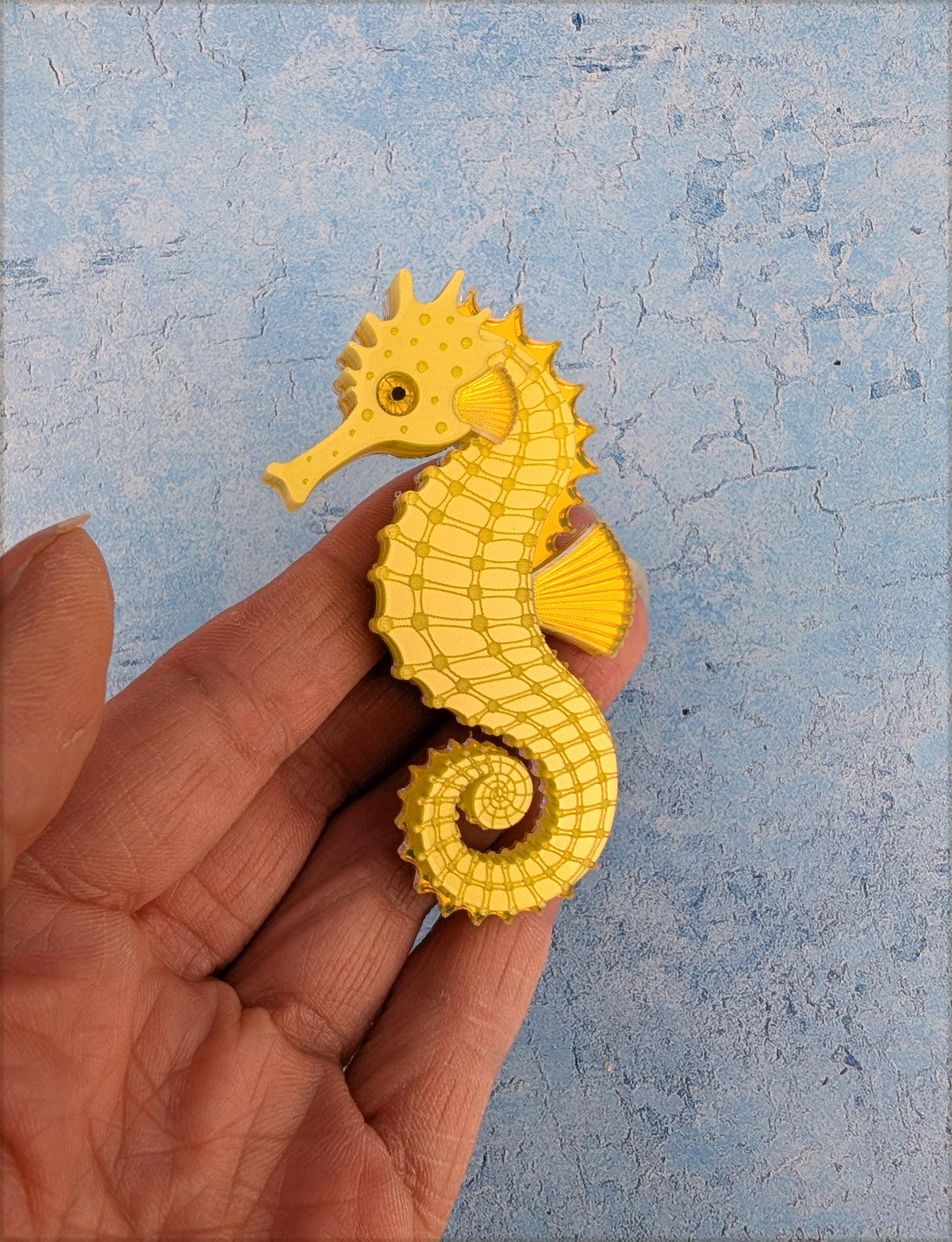 A small yellow acrylic seahorse
 brooch held in a person's hand against a textured light blue background.