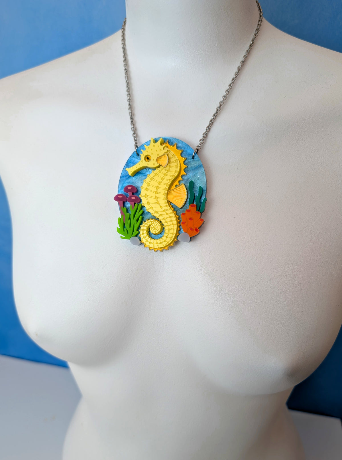 A silver chain necklace with an oval pendant featuring a yellow seahorse and colorful sea life details, displayed on a white mannequin torso against a blue background.