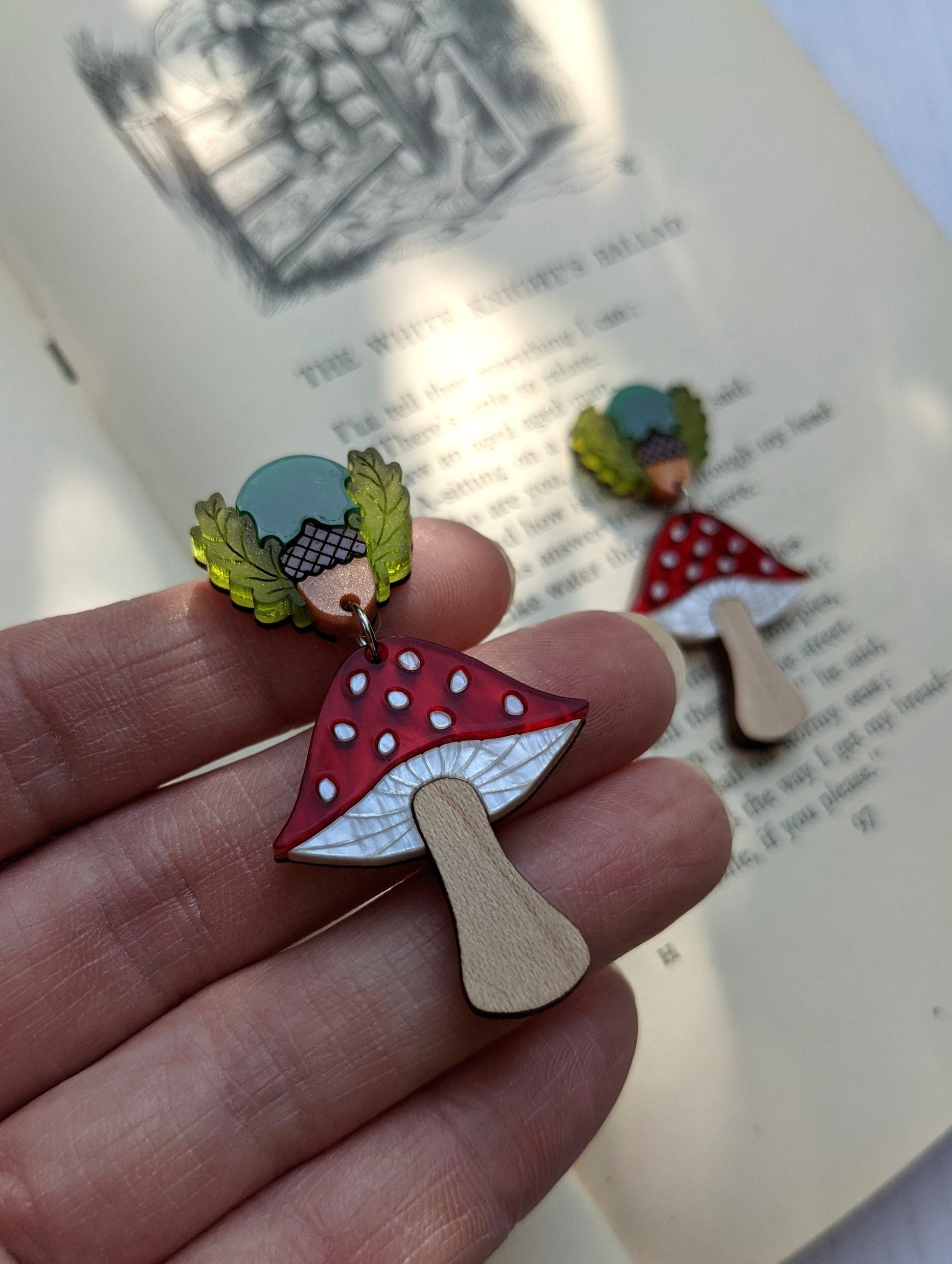 A person holding laser cut earrings in the shape of toadstools with caps in red, green, yellow and brown, stem in light beige, attached to butterfly backs.