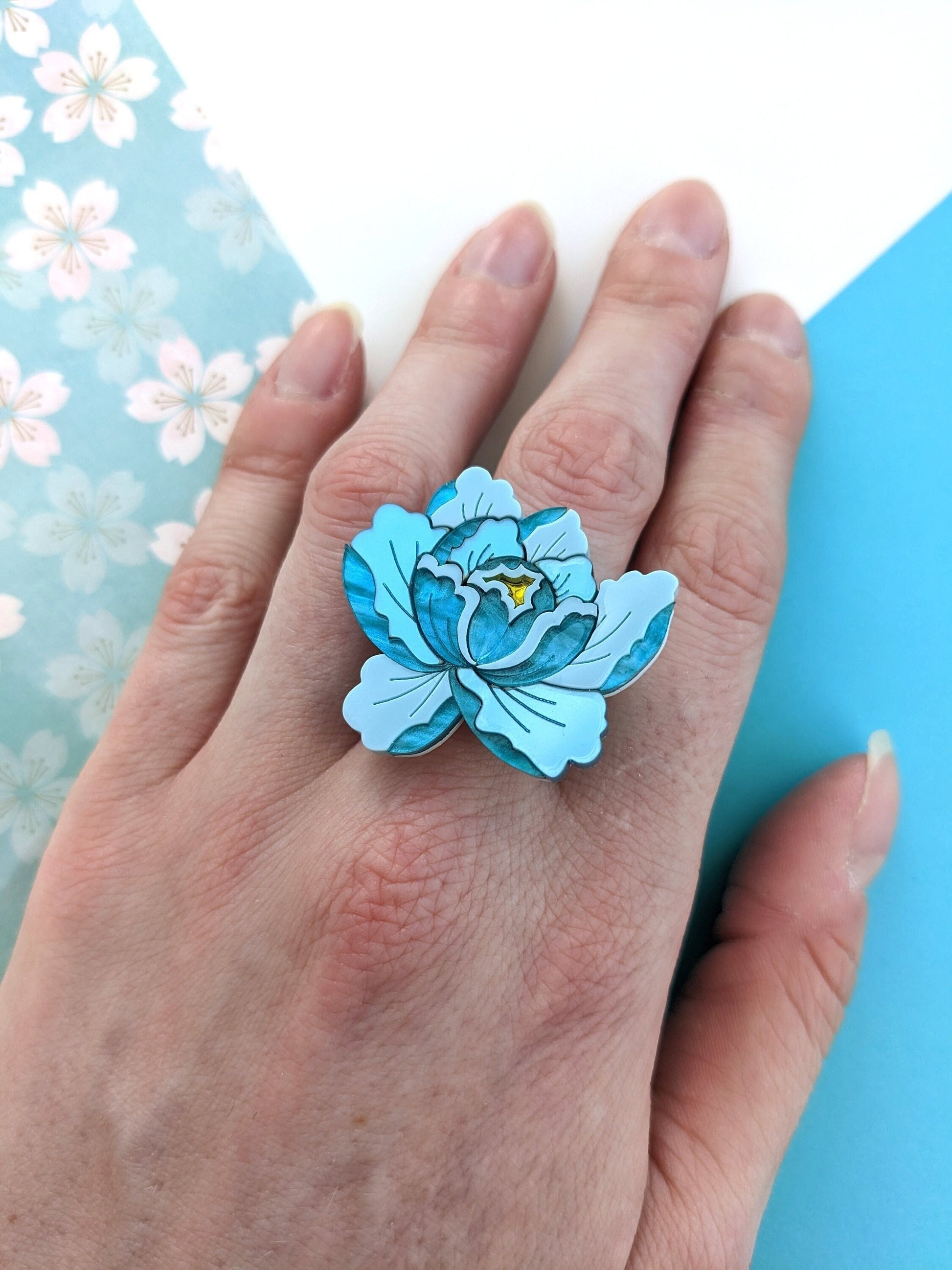 A hand wearing a teal floral ring with adjustable band, made of laser-cut acrylic pieces.