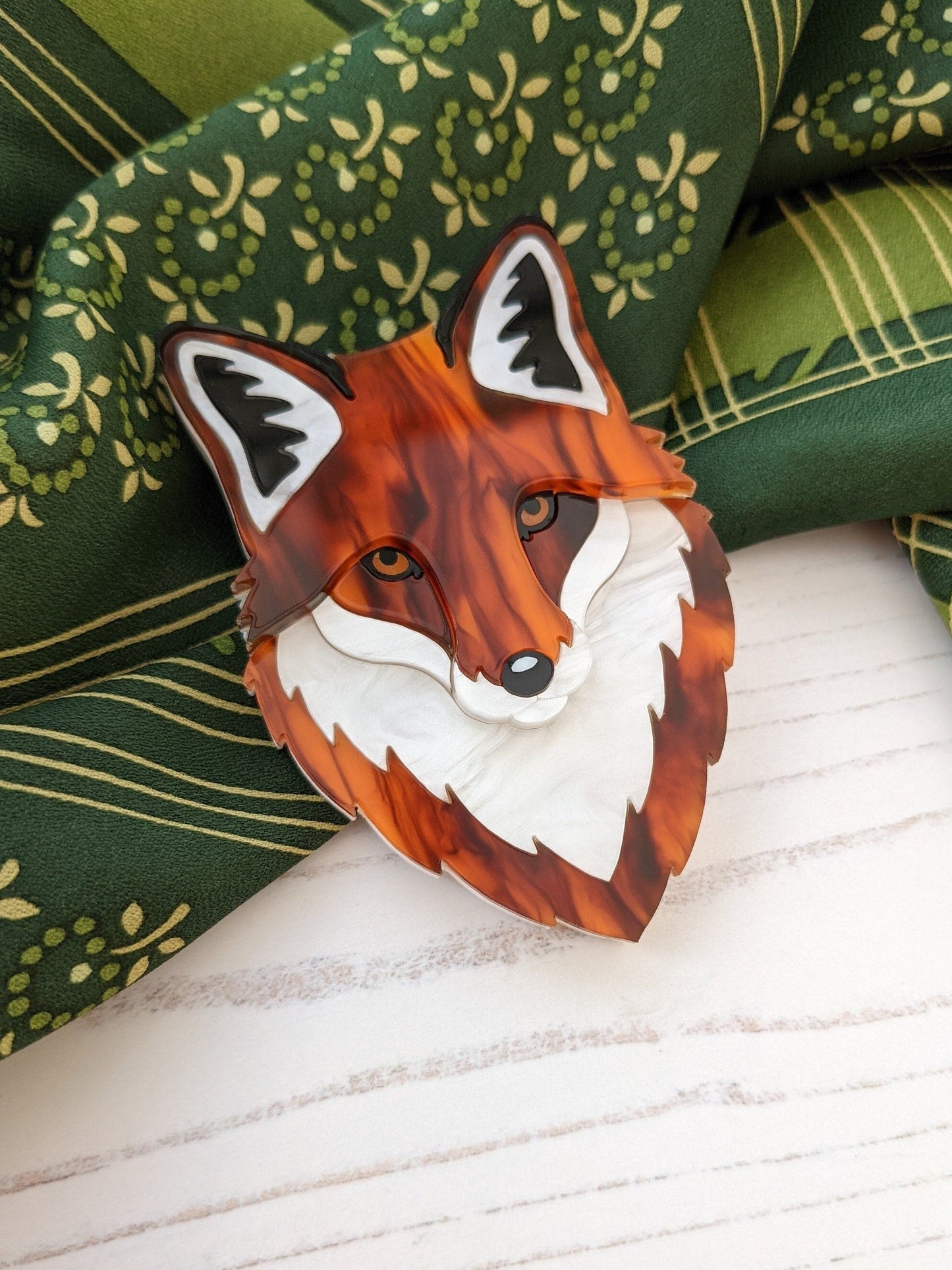 A laser cut acrylic brooch in the shape of a fox head, featuring marbled pearl white acrylic with black and blue accents, displayed on a green patterned fabric.