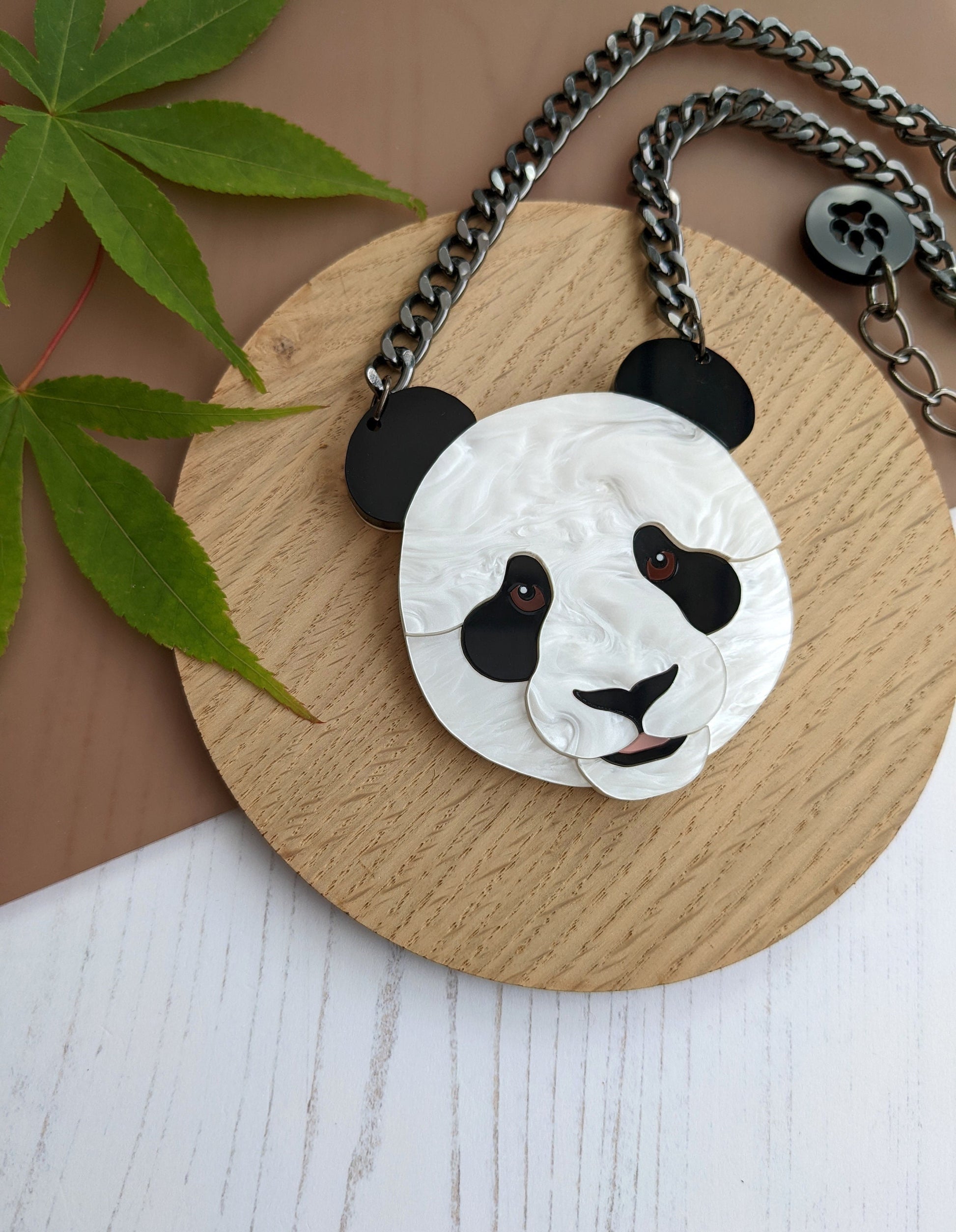 A necklace with a 3D acrylic panda head pendant on a gunmetal colored chain, displayed on a wooden round tray with a leaf to the side.