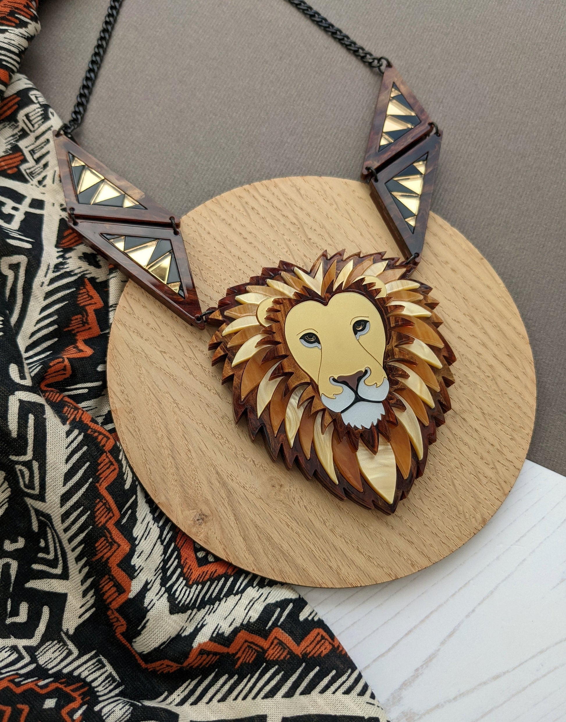 A statement necklace featuring a lion design made from gold and caramel colored acrylics, with black details and pearl-like accents, suspended from a black chain.
