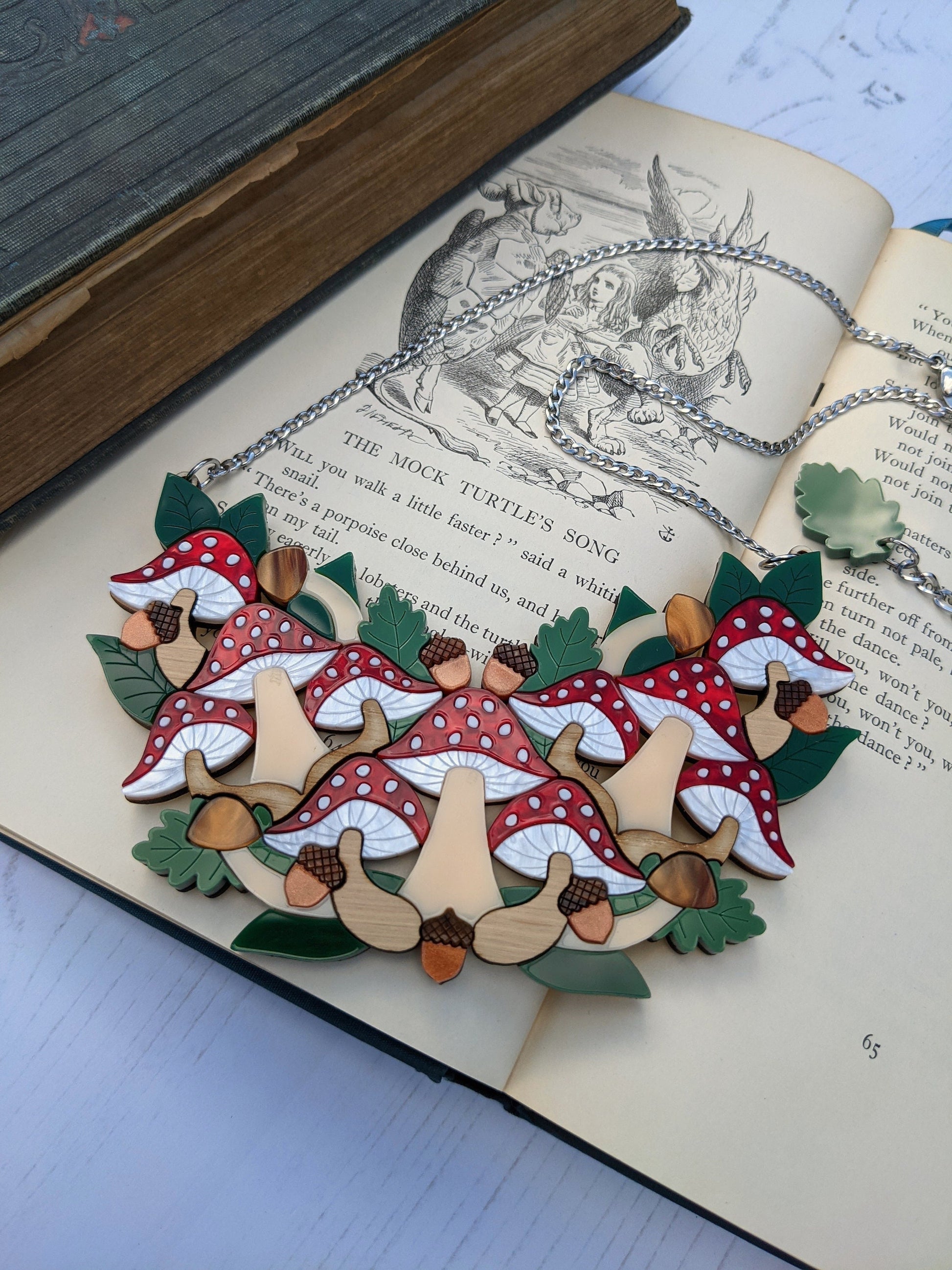 A toadstool statement bib necklace displayed on an open book with illustrations related to the product.