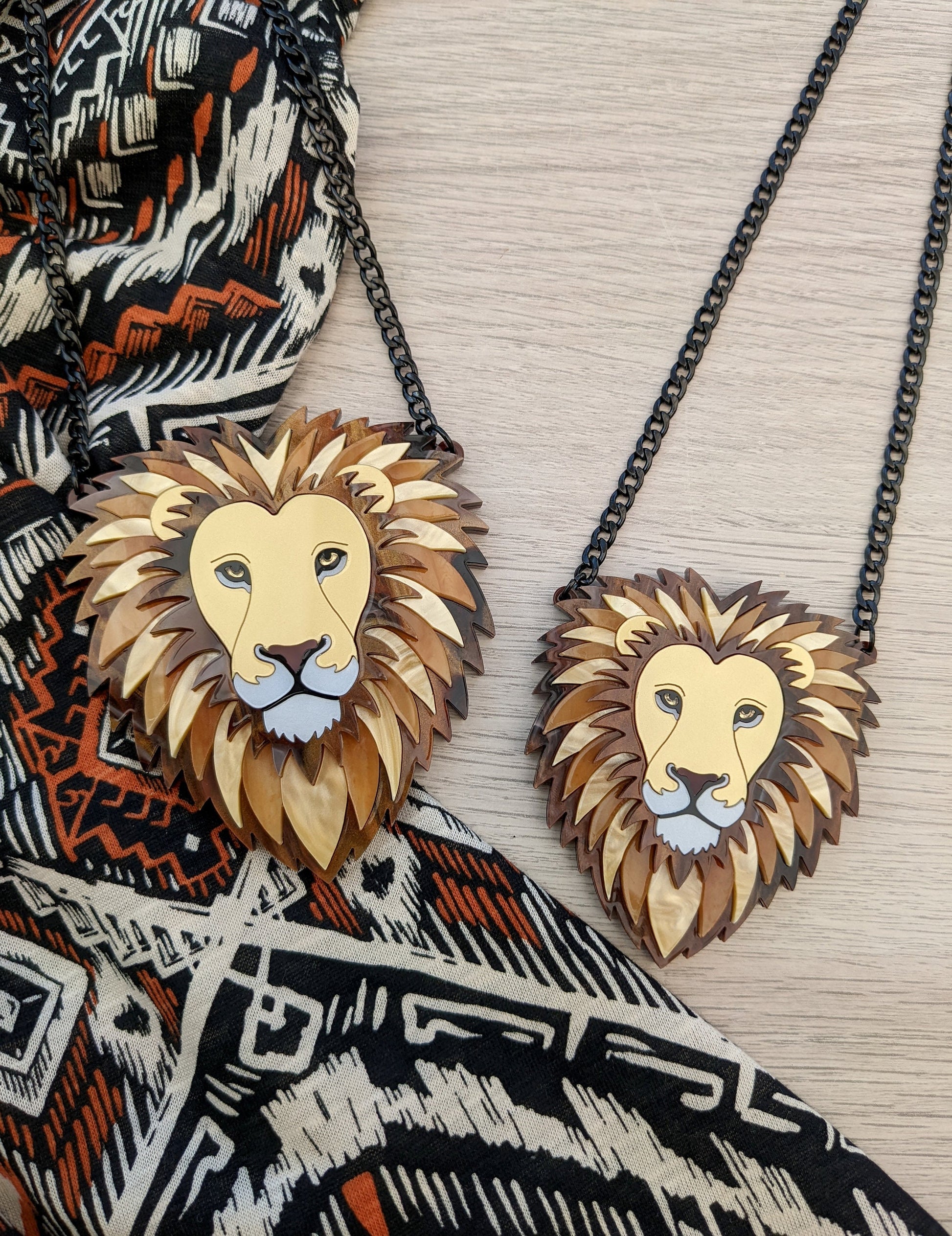 Two acrylic lion necklaces on display, one larger and one smaller, with a black chain and a paw charm. The necklaces have gold and caramel colored details, and are placed against a patterned backdrop.